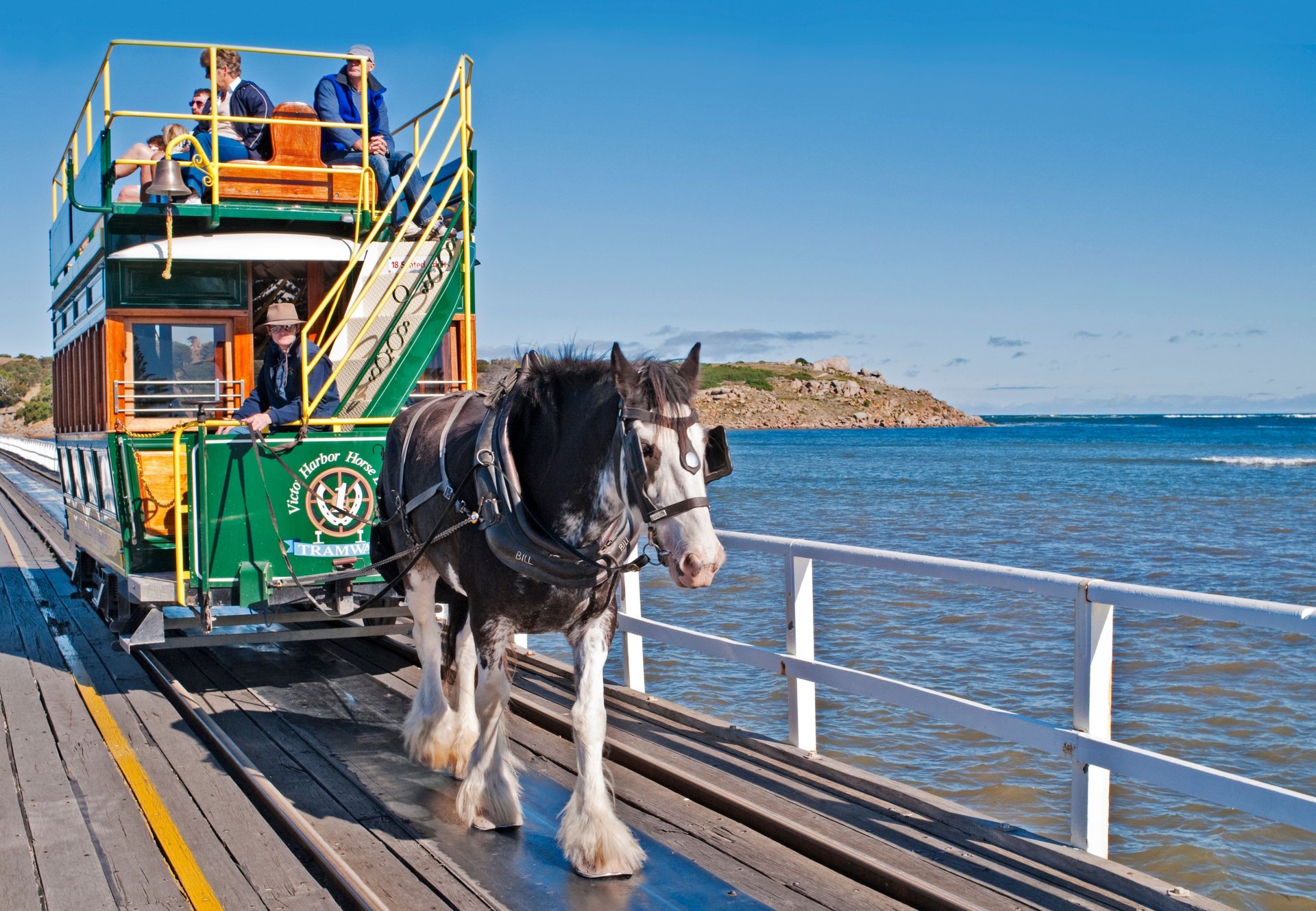A horse-drawn tram crosses a wooden pier over water, with passengers seated on the upper deck under a clear blue sky.