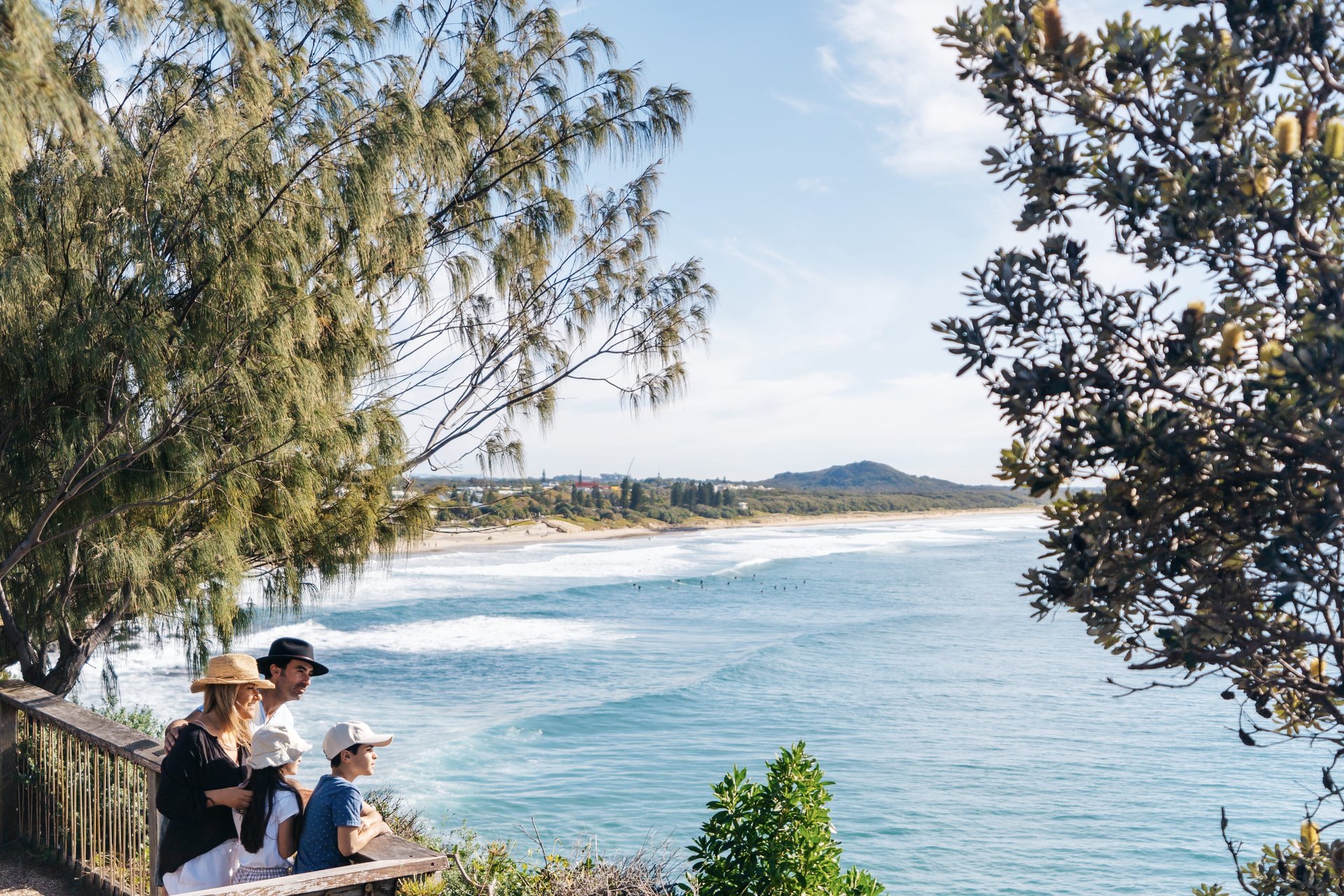 A family stands on a wooden deck overlooking a scenic coastal view with waves, trees, and distant hills under a clear blue sky.
