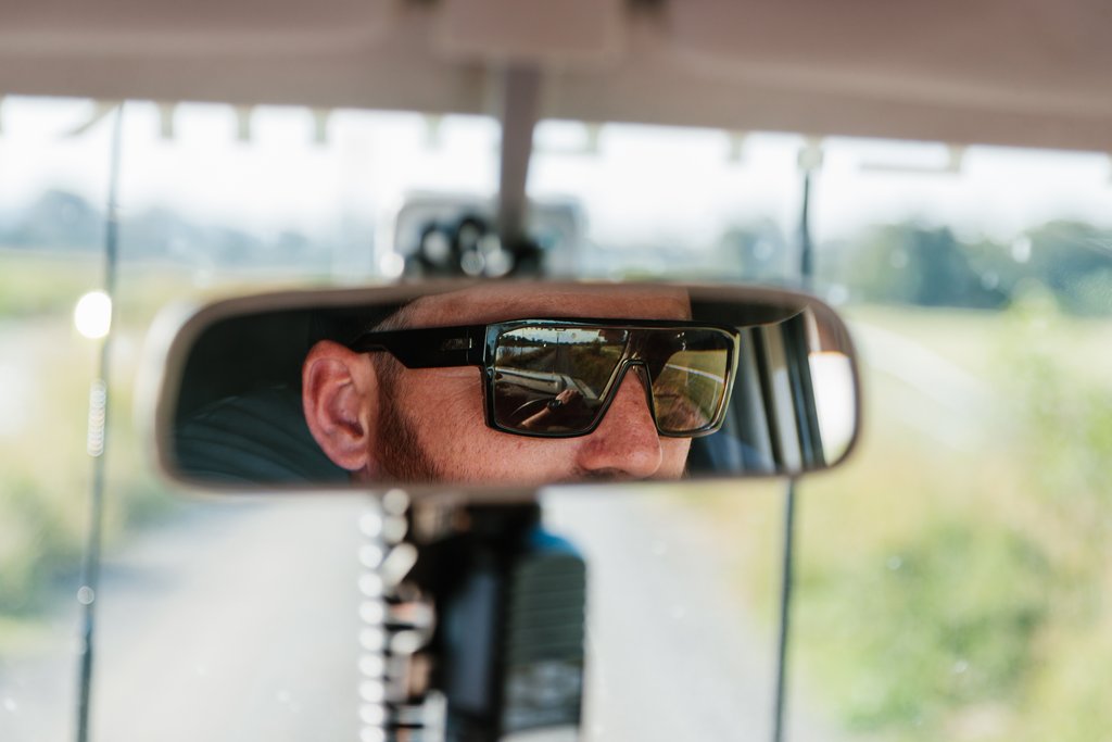 Reflection of a person wearing sunglasses, visible in a car's rearview mirror, with a blurred outdoor scene in the background.