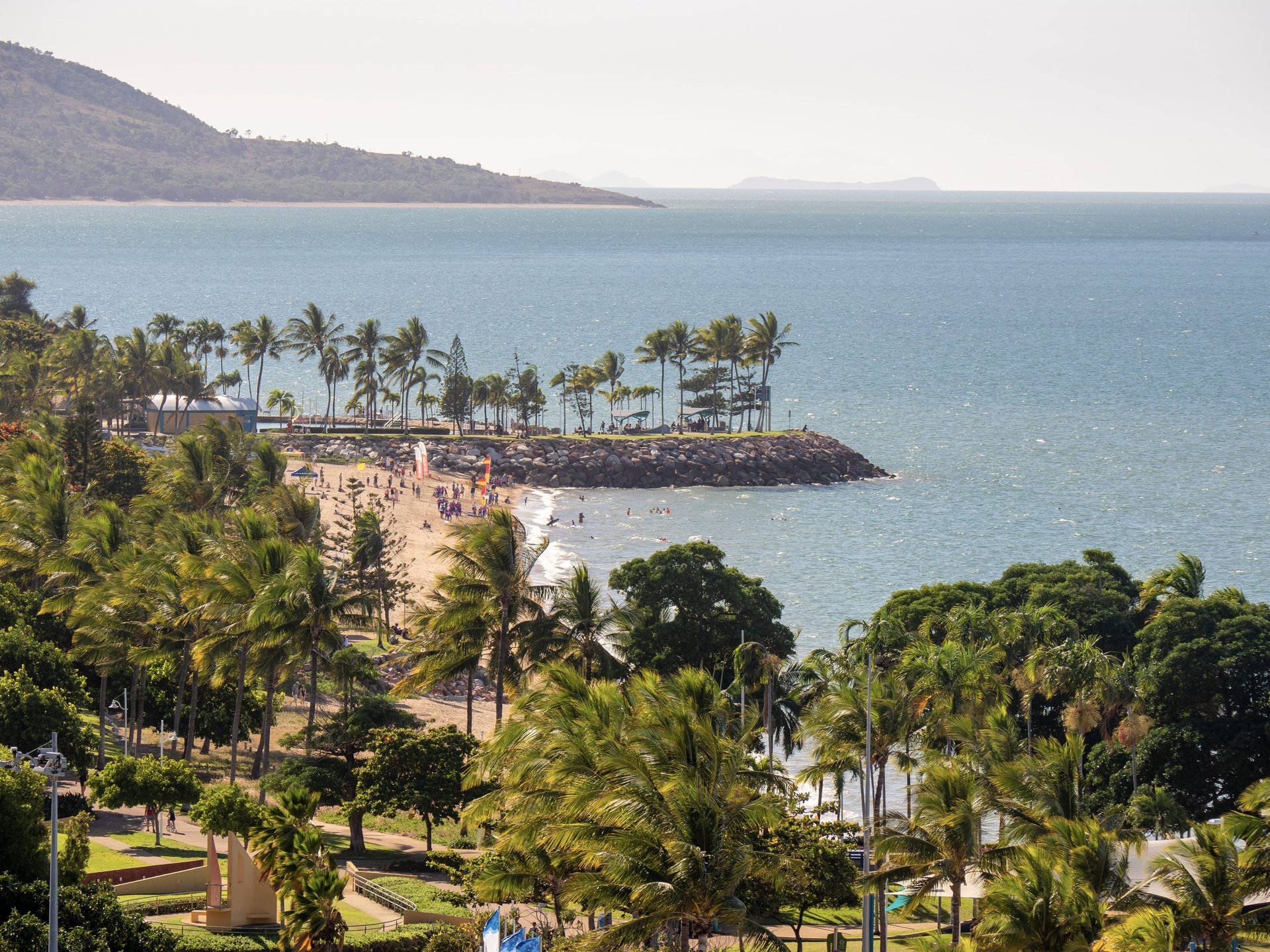 A sandy tropical beach lined with palm trees, people onshore, rocky breakwater jutting into calm blue sea and distant hills.