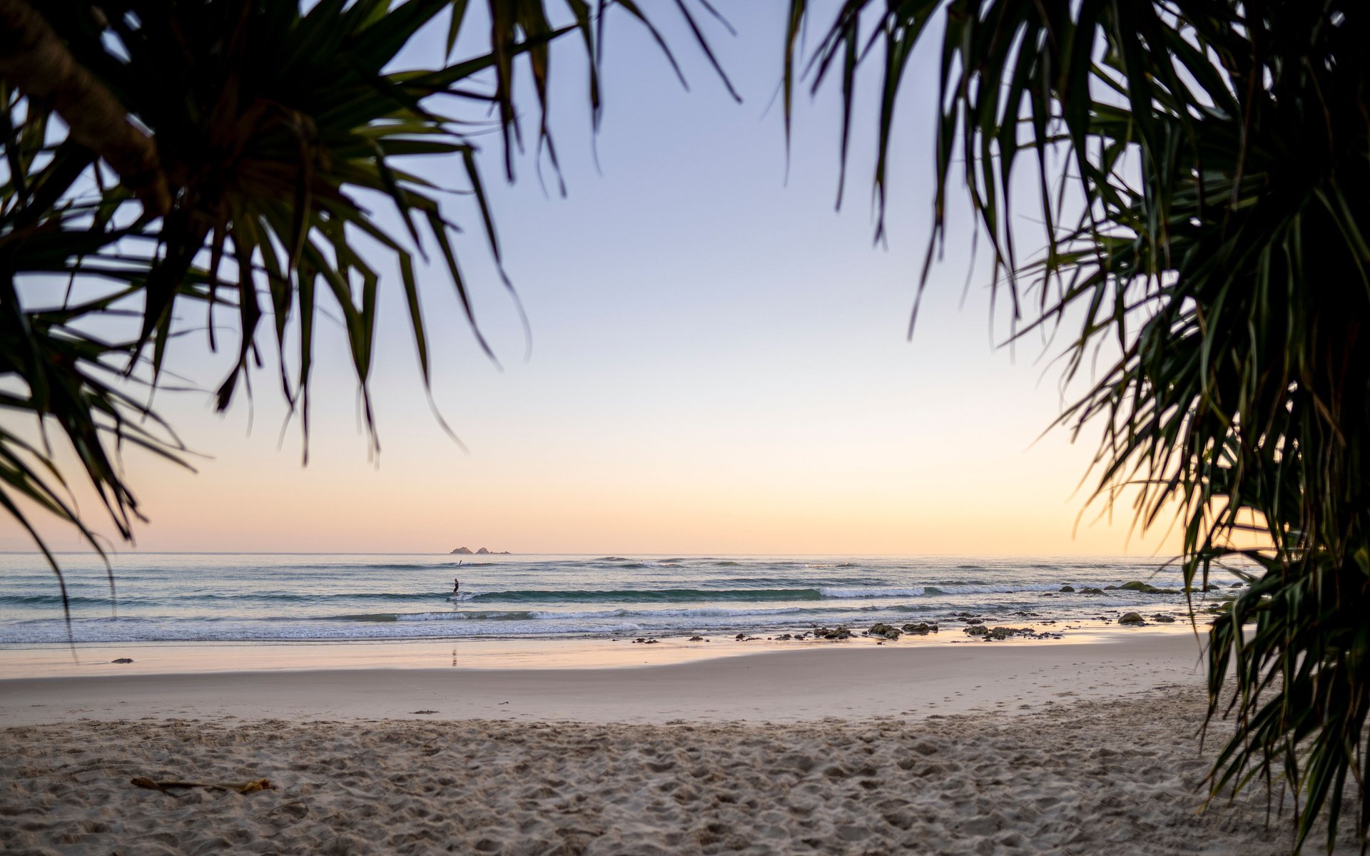 Sunrise beach framed by pandanus leaves, calm waves, lone paddleboarder and distant rocky islets on the horizon.