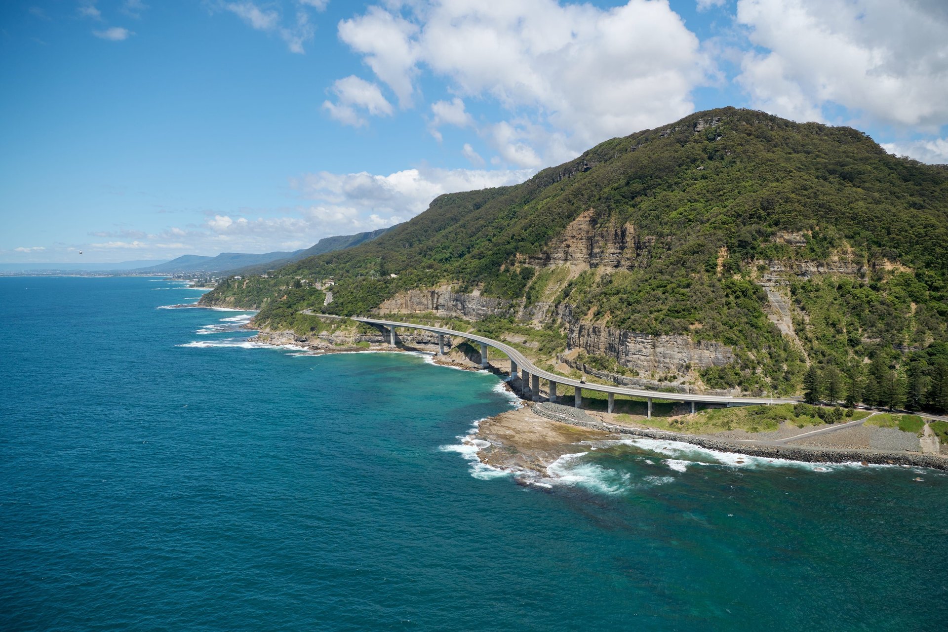 Curved coastal bridge hugging forested cliffs above turquoise sea, waves breaking on the rocky shoreline under a blue sky