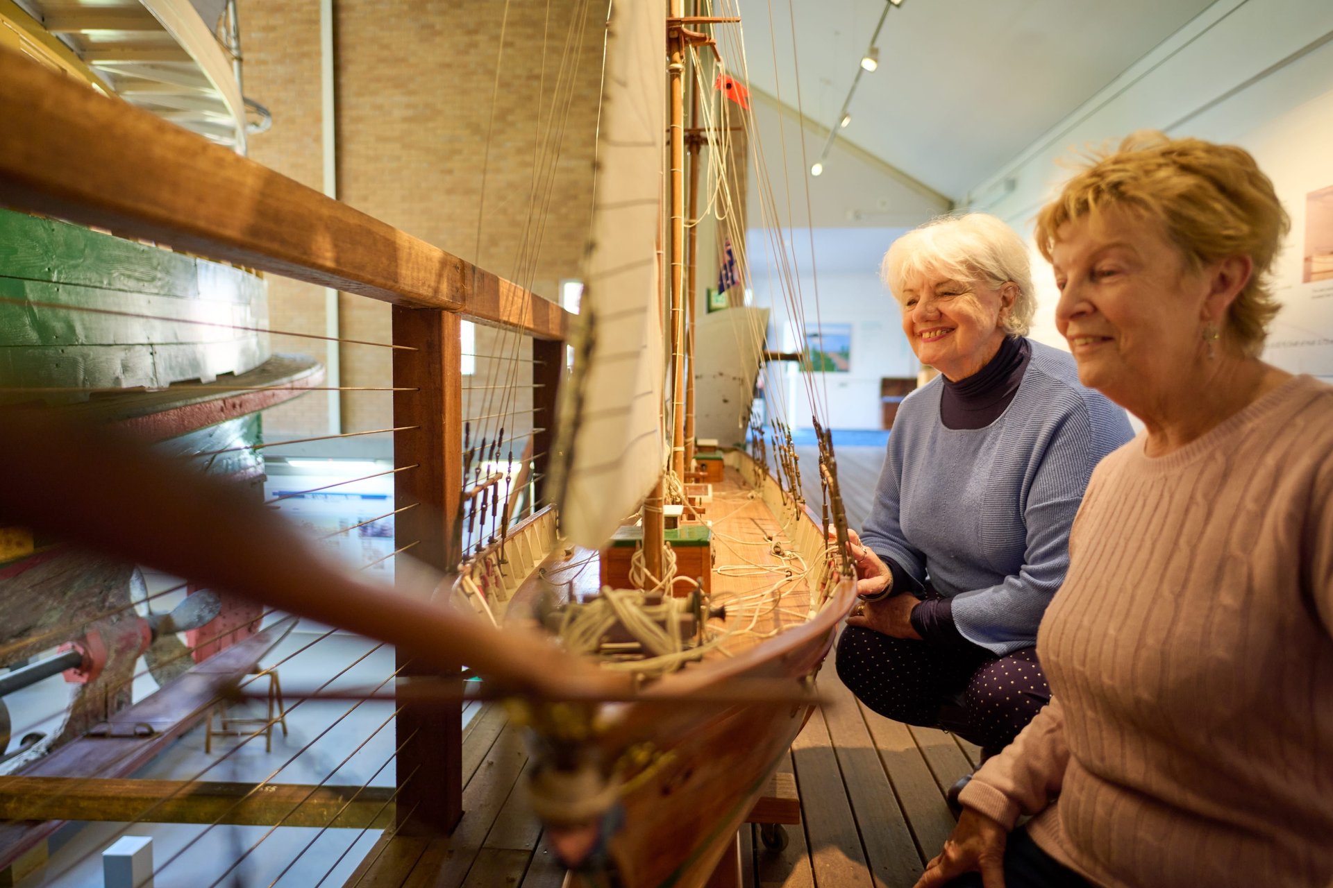 Two older women smiling as they examine a detailed wooden model sailboat in a museum.