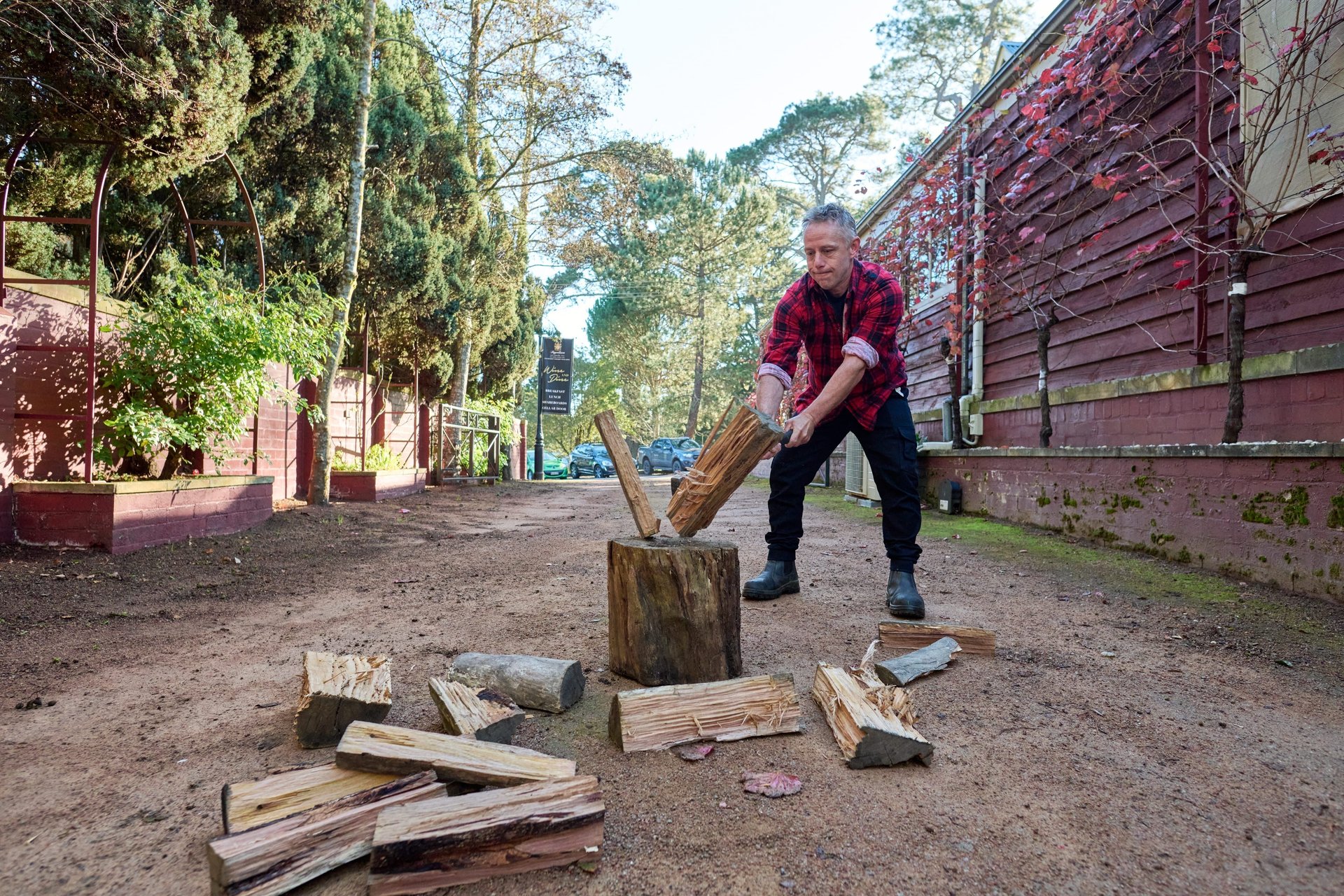 Man in red plaid shirt splitting firewood on a stump in a dirt lane beside a red wooden building, scattered logs around.