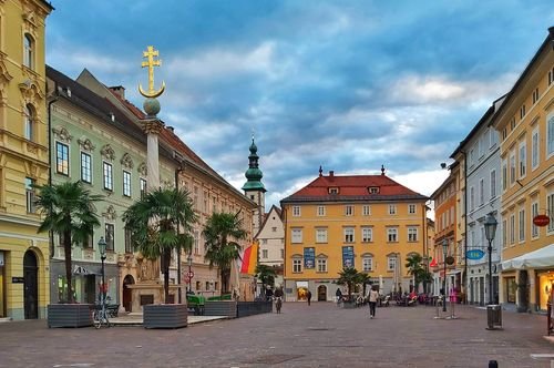 Ein malerischer europäischer Platz mit bunten historischen Gebäuden, Palmen und einem bewölkten Himmel. Menschen gehen spazieren und fahren Fahrrad.