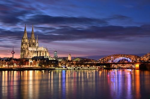 Der Kölner Dom und die Hohenzollernbrücke, beleuchtet in der Dämmerung, spiegeln sich im Rhein unter einem lebendigen Himmel wider.