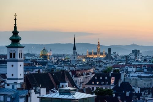 Ein Panorama einer Stadtlandschaft in der Dämmerung mit historischen Gebäuden, Türmen und einer sanft beleuchteten Skyline unter einem farbverlaufenden Abendhimmel.