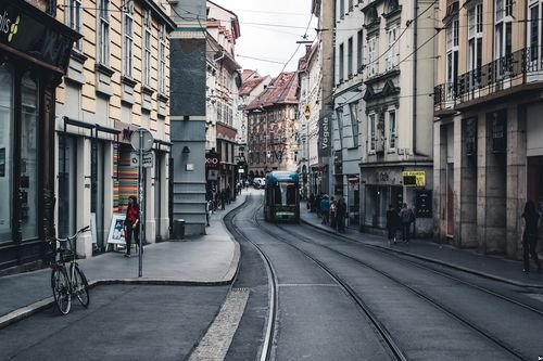 Schmale europäische Straße mit einer Straßenbahn auf Gleisen, umgeben von historischen Gebäuden und Geschäften; Fahrräder sind auf dem Gehweg geparkt.