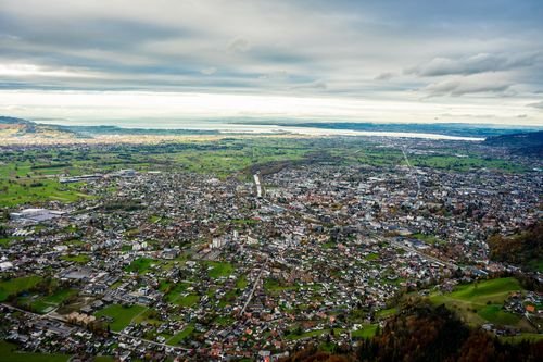 Luftaufnahme einer ausgedehnten Stadt mit dichter Bebauung, umgeben von grünen Feldern und entferntem Wasser unter einem bewölkten Himmel.