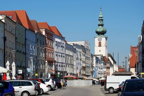 Eine Straße gesäumt von bunten historischen Gebäuden und parkenden Autos, die zu einer hohen Kirche mit grüner Kuppel unter einem klaren blauen Himmel führt.