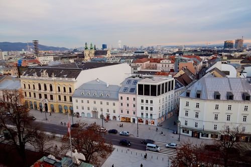 Luftaufnahme einer europäischen Stadtlandschaft mit historischen Gebäuden, einer belebten Straße und entfernten modernen Wolkenkratzern unter einem bewölkten Himmel.