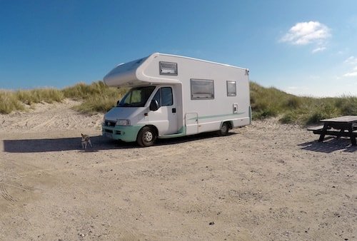 A white camper van parked on a sandy beach near grassy dunes under a clear blue sky, with a small dog standing nearby.