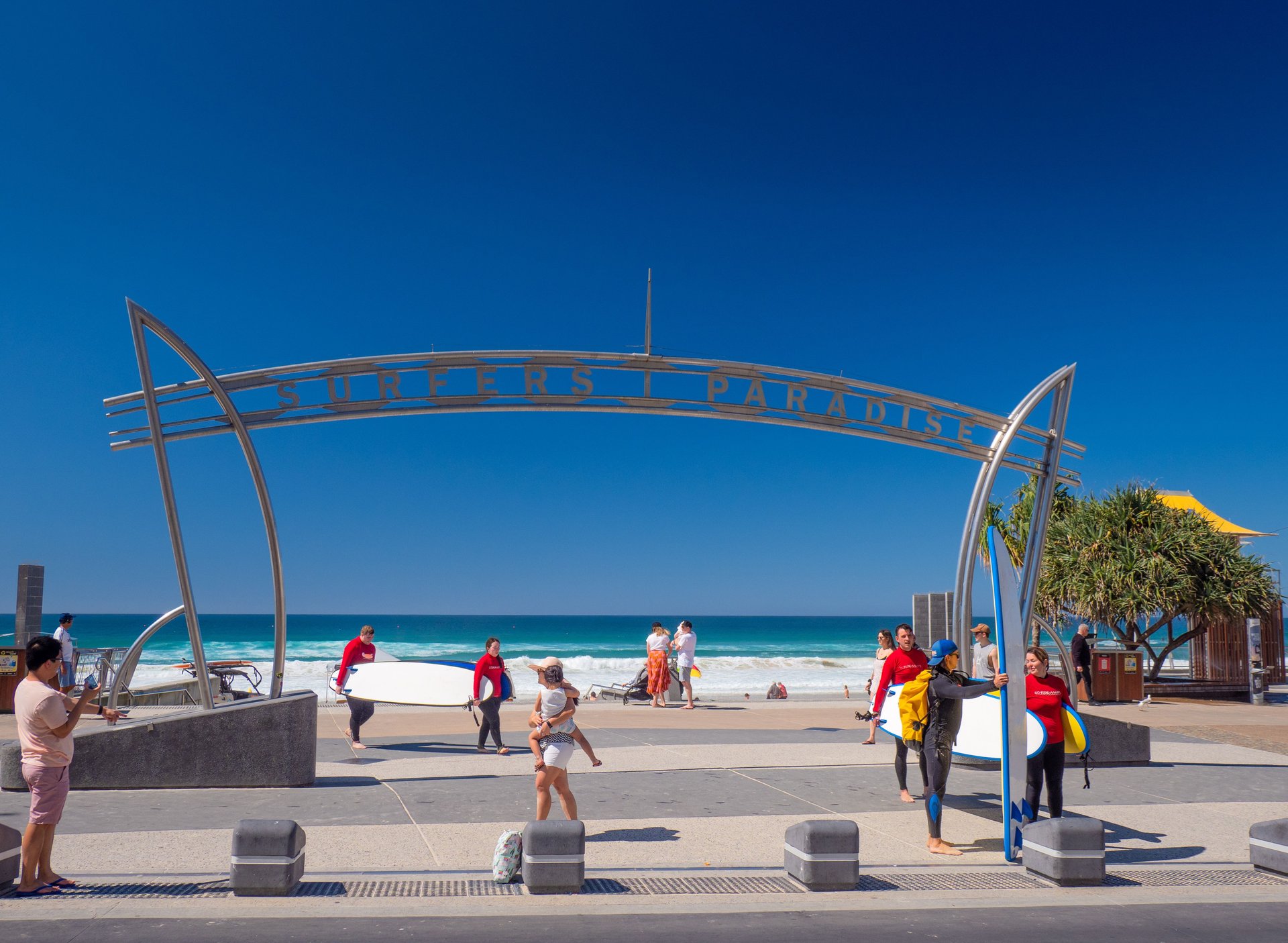 People with surfboards walk under the "Surfers Paradise" sign by a sunny beach, with a clear blue sky and ocean waves in the background.
