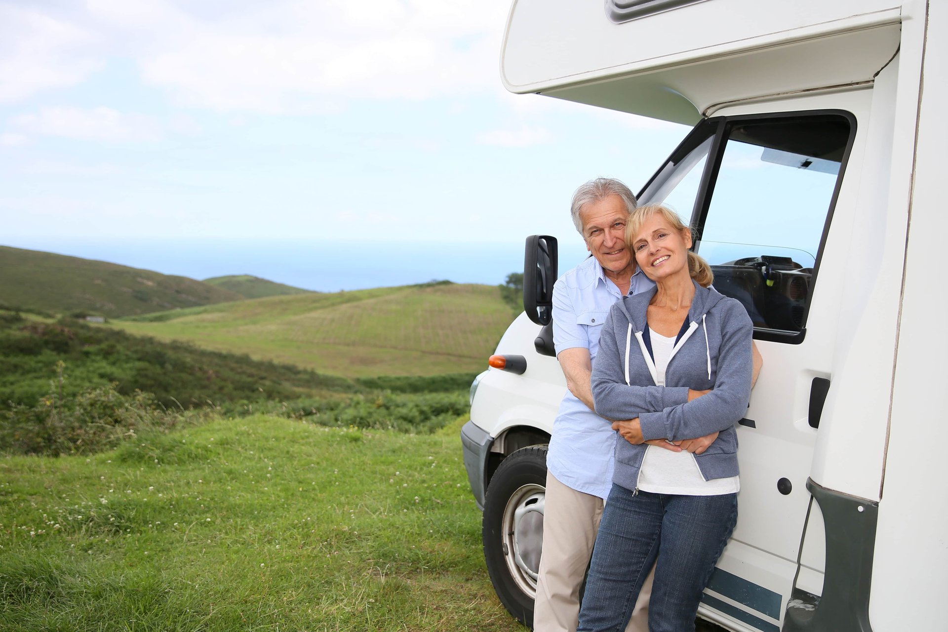 Older couple smiling and embracing by a camper van on a grassy hill, with a scenic view of rolling hills and the ocean in the background.