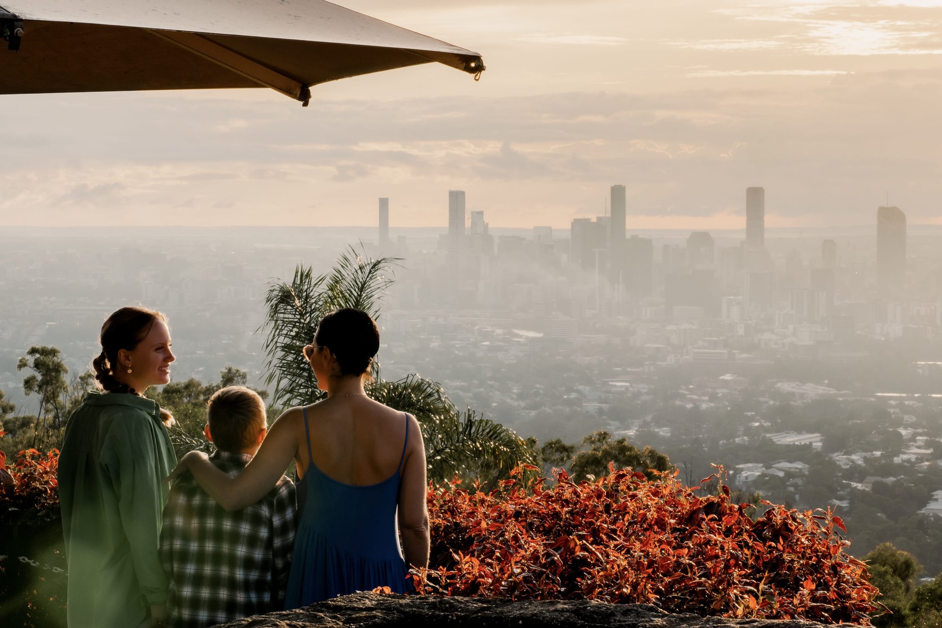 Three people overlook a city skyline at sunset, with lush foliage and a sunlit sky creating a serene atmosphere.