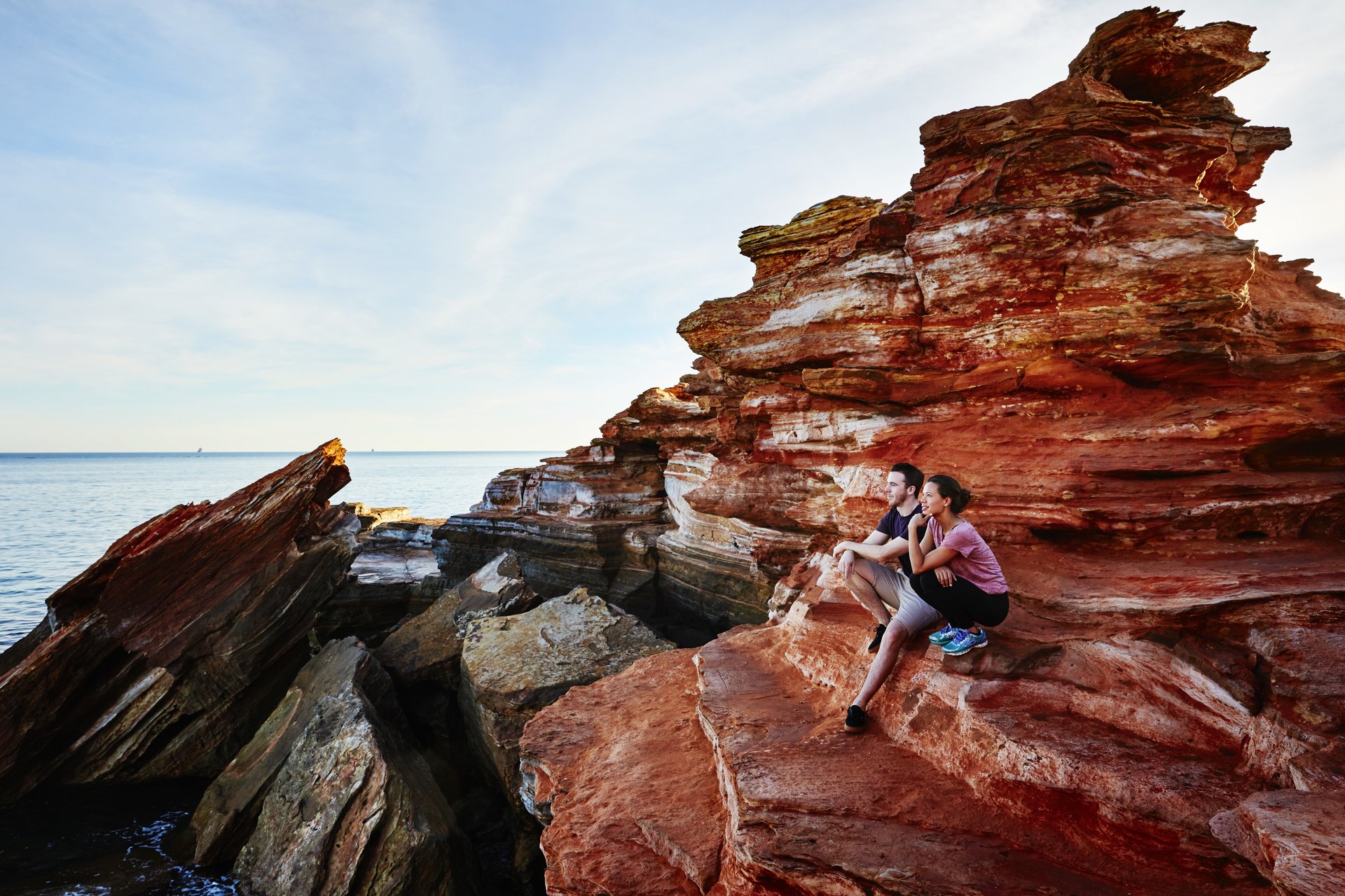 Couple seated on layered red sandstone cliffs, gazing out over a calm sea beneath a wide blue sky.
