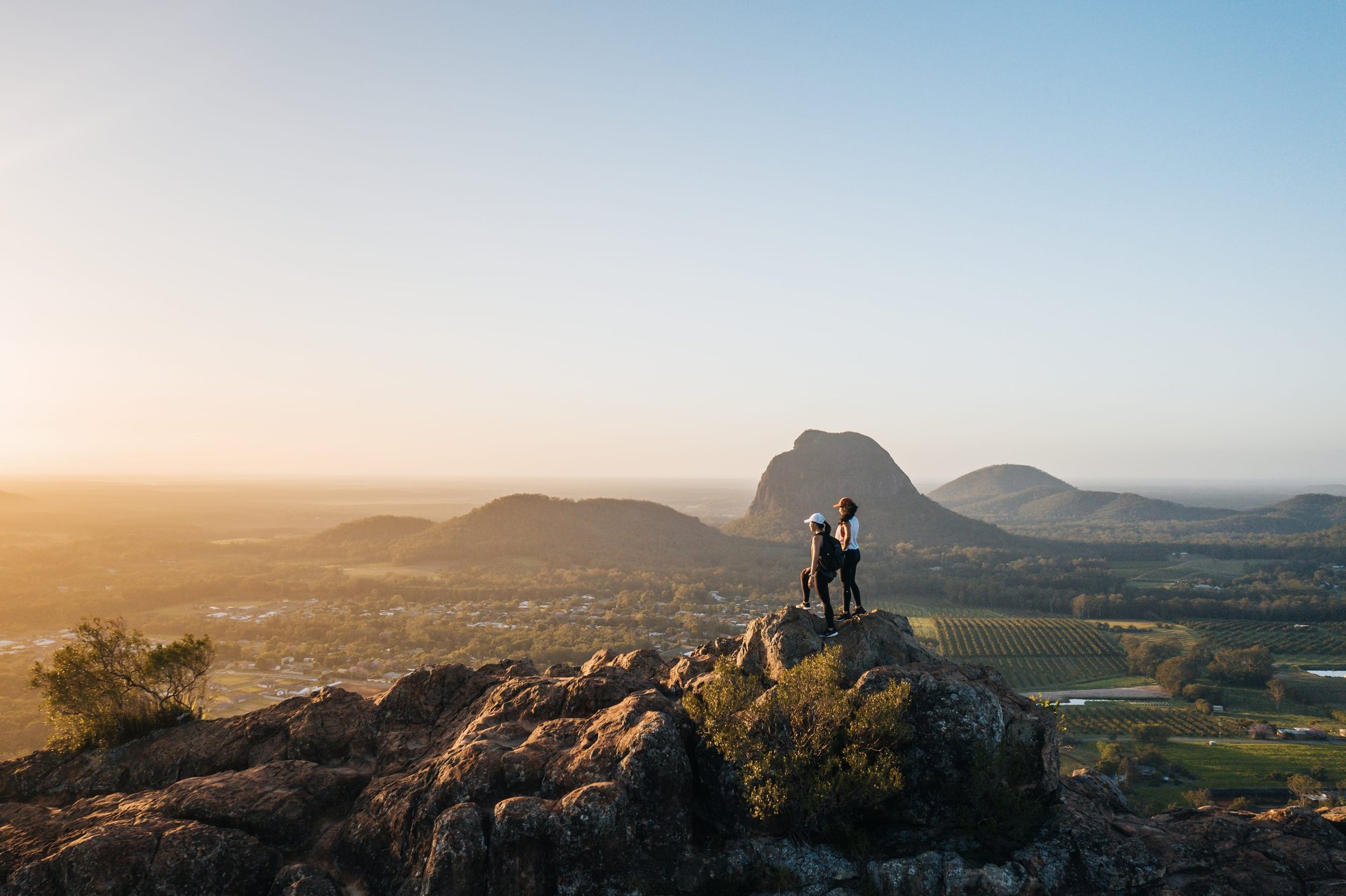 Two people stand on a rocky peak, overlooking a vast landscape with fields and distant hills under a clear sky at sunset.
