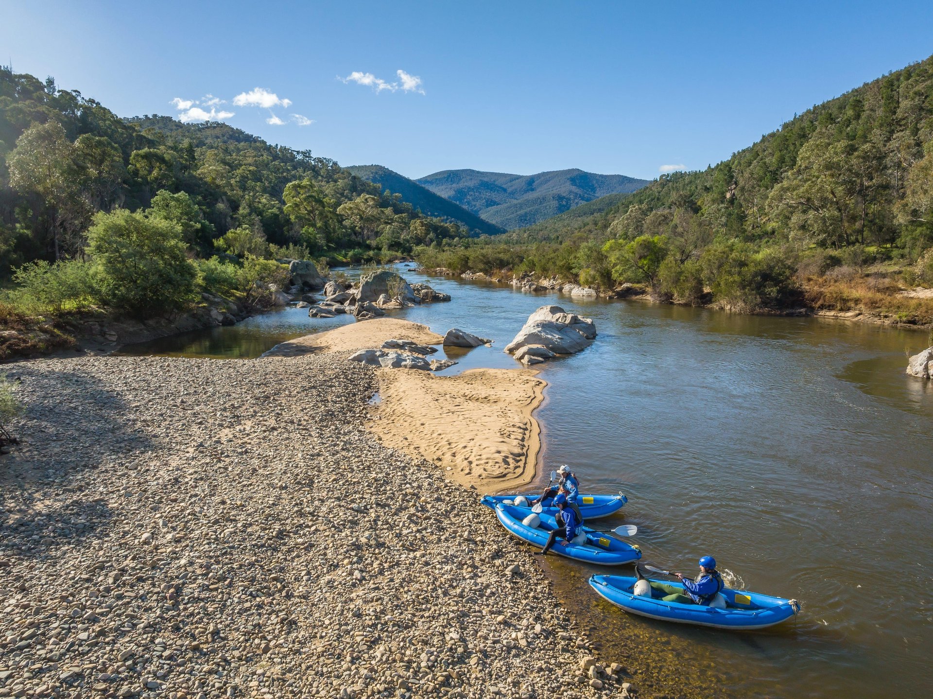 Three blue inflatable kayaks with paddlers on a river beside a rocky shore and sandbar, forested hills and clear sky.