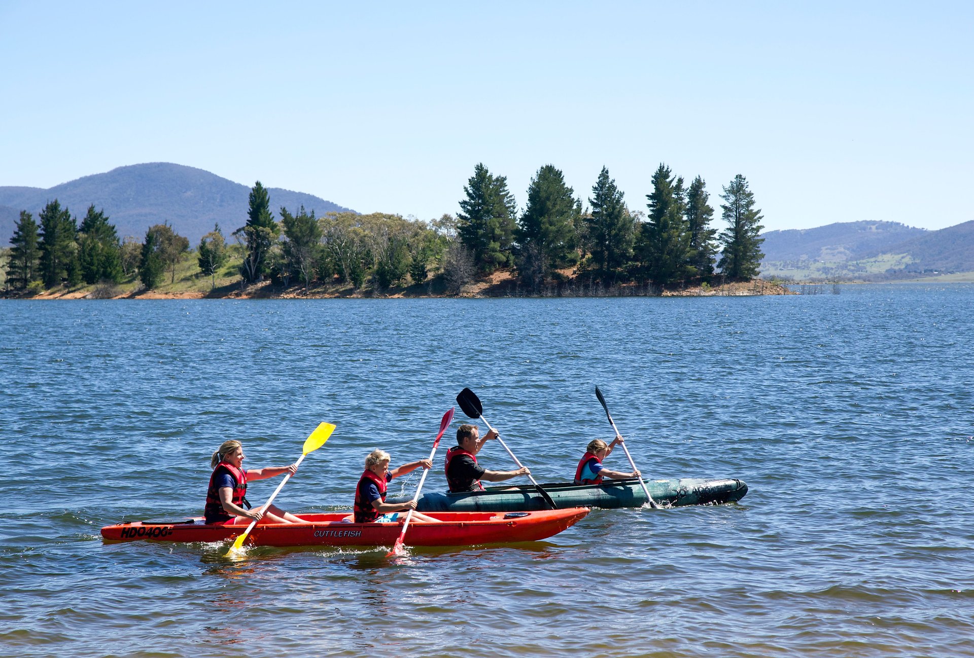 Four people wearing life jackets in two kayaks paddling on a lake with a tree-lined island and distant hills under a clear sky.