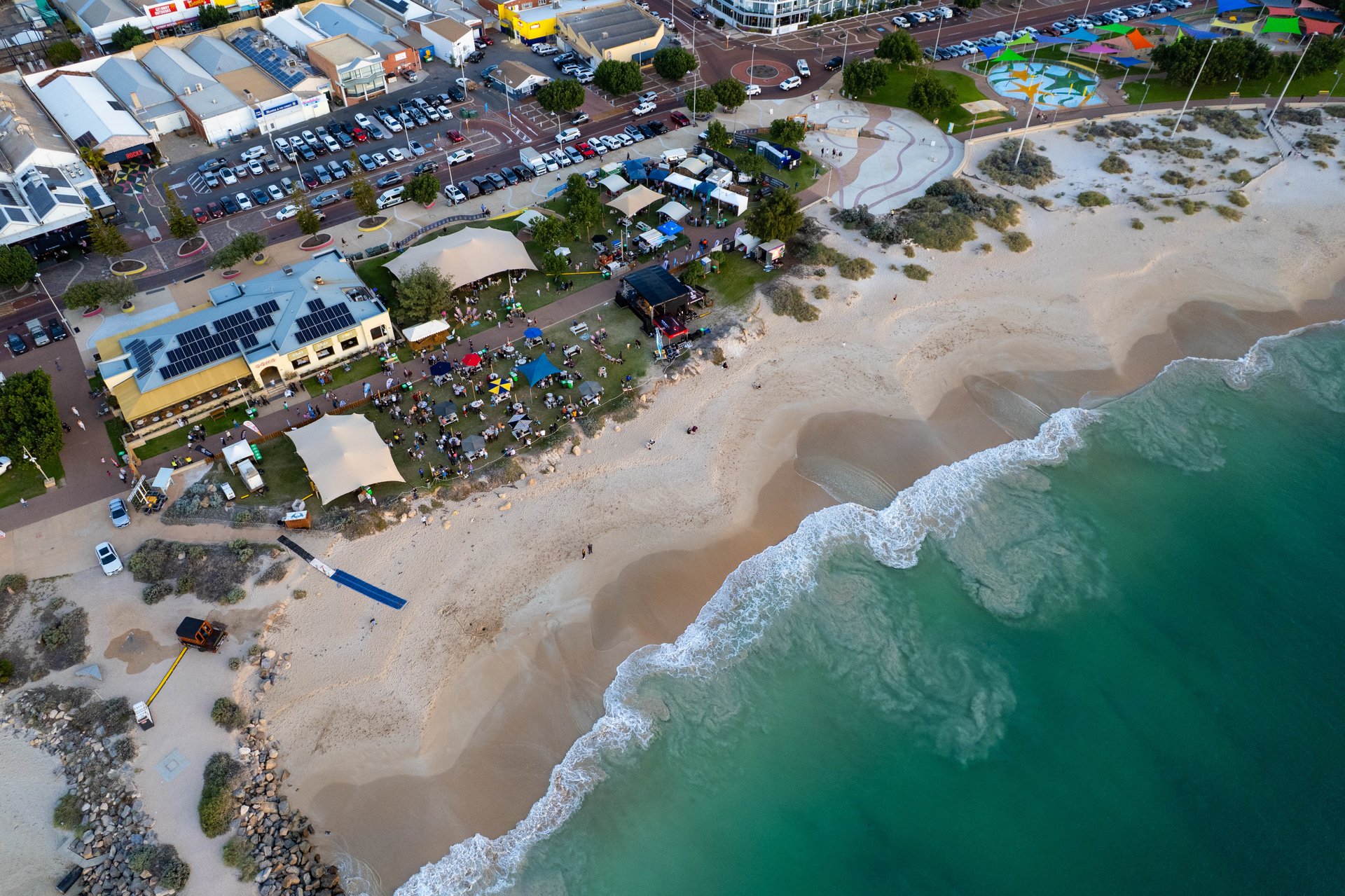 Aerial view of a beachfront festival: tents and crowds on sand dunes, parked cars, promenade and turquoise waves.