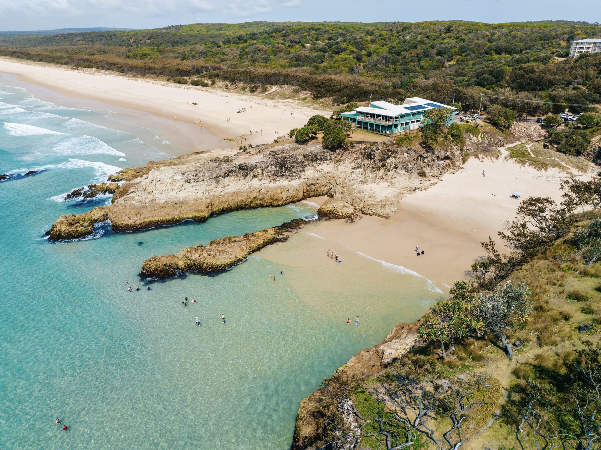 Aerial view of a coastal beach with turquoise waters, rocky outcrops, and a building surrounded by greenery on a sunny day.