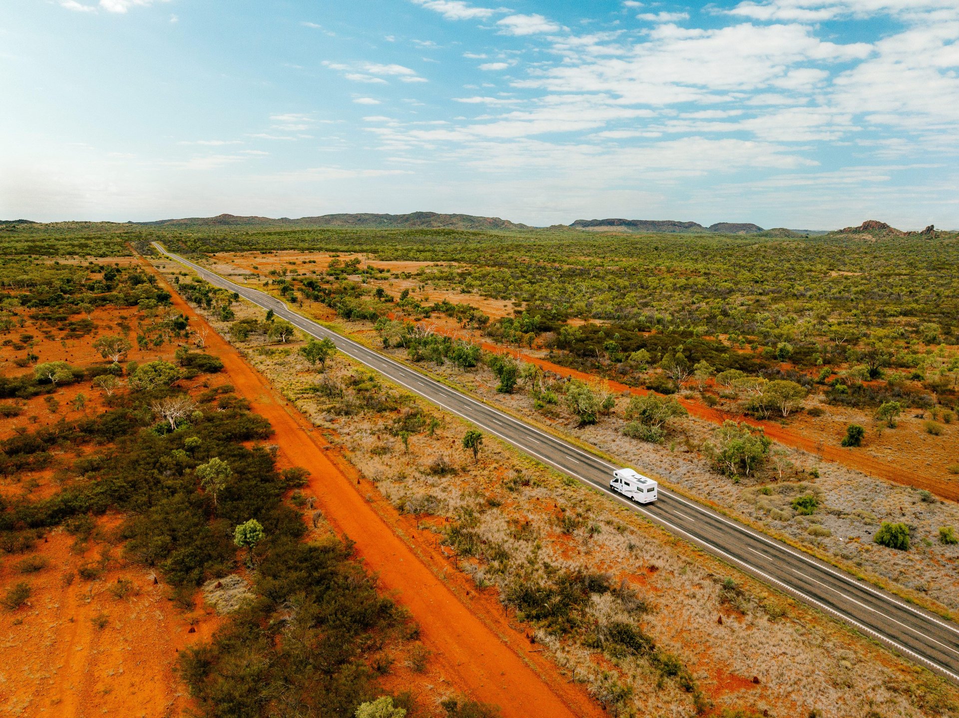 Aerial view of a white RV traveling on a long, straight road through a vast, arid landscape with red soil and sparse greenery under a blue sky.