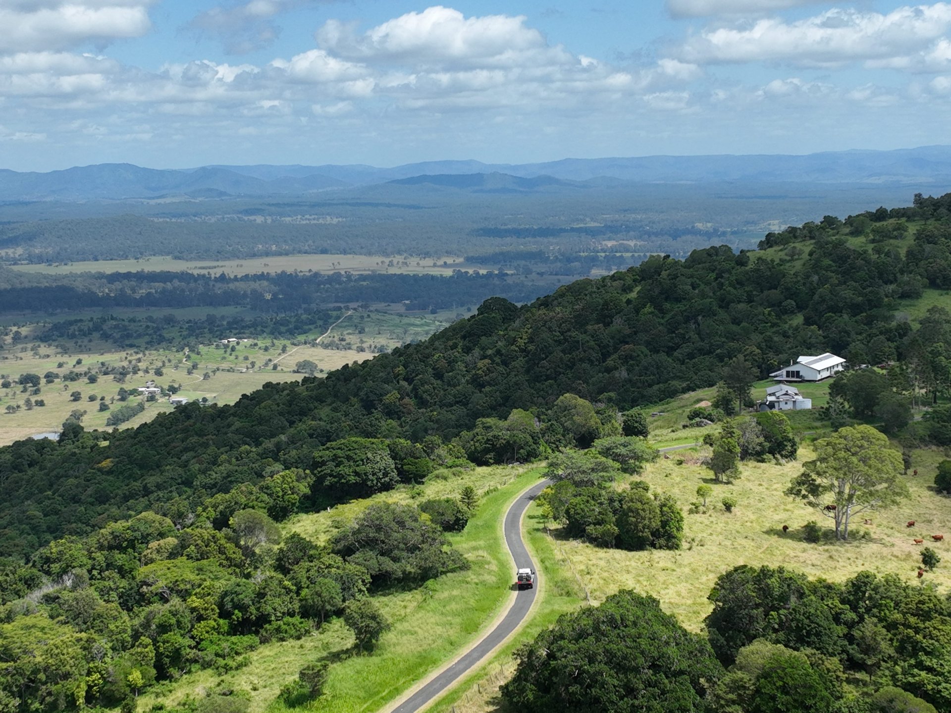 Winding country road with a small car through lush green hills, scattered houses, a broad valley and distant blue mountains.
