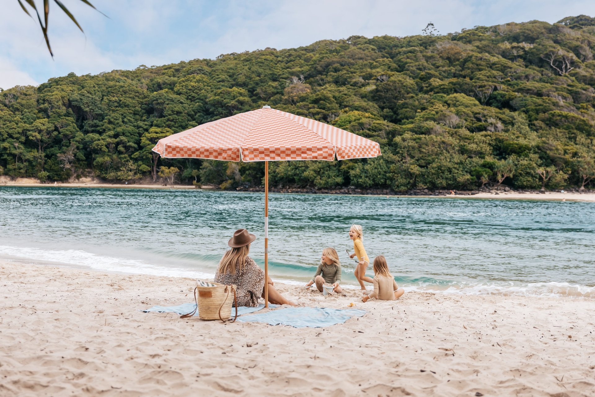 A woman and three children sit on a sandy beach under a red-and-white umbrella, with a forested hill in the background.