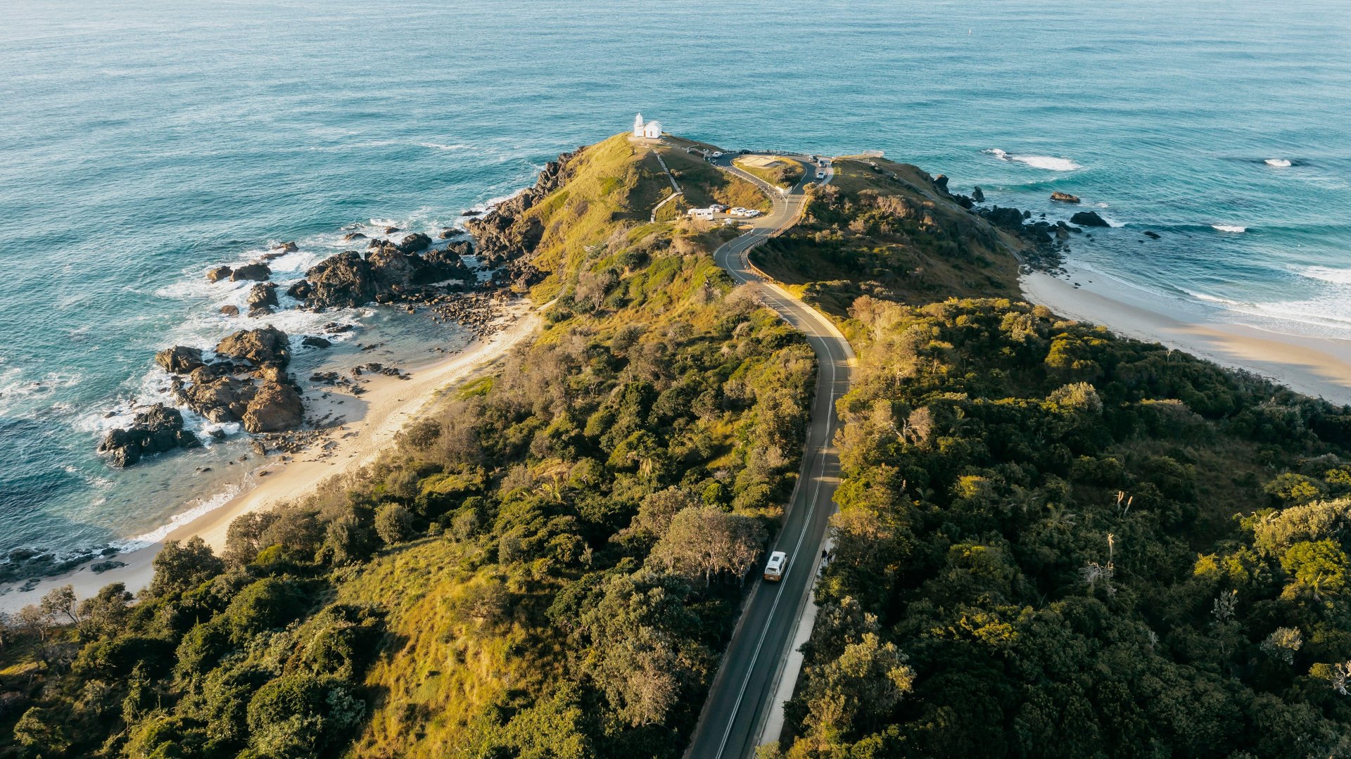 Aerial view of a winding coastal road with a camper van leading to a white lighthouse on a green headland, rocky beach and blue sea.