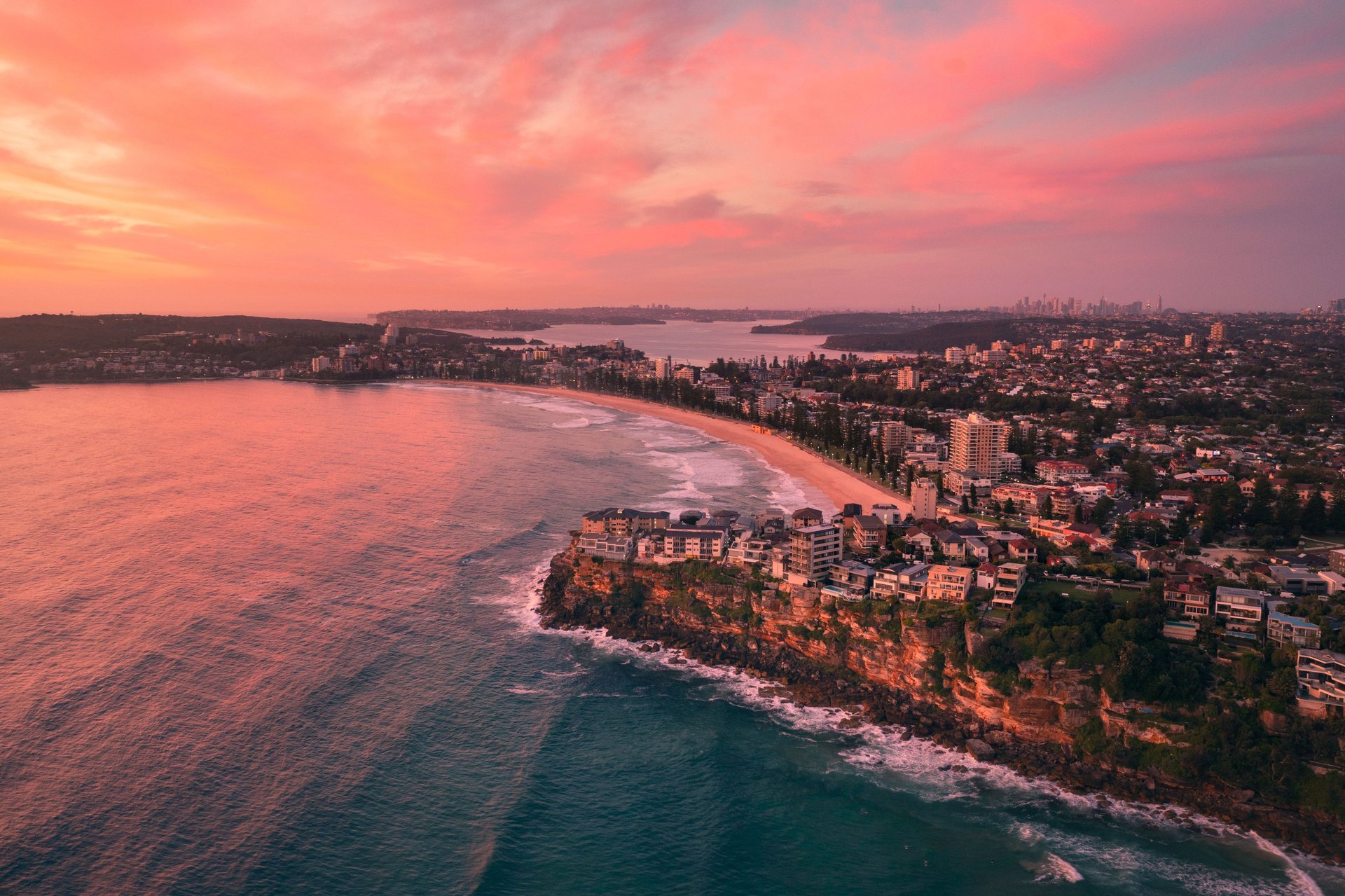 Aerial view of a coastal city at sunset, with pink skies, a sandy beach, and waves crashing against rocky cliffs lined with buildings.