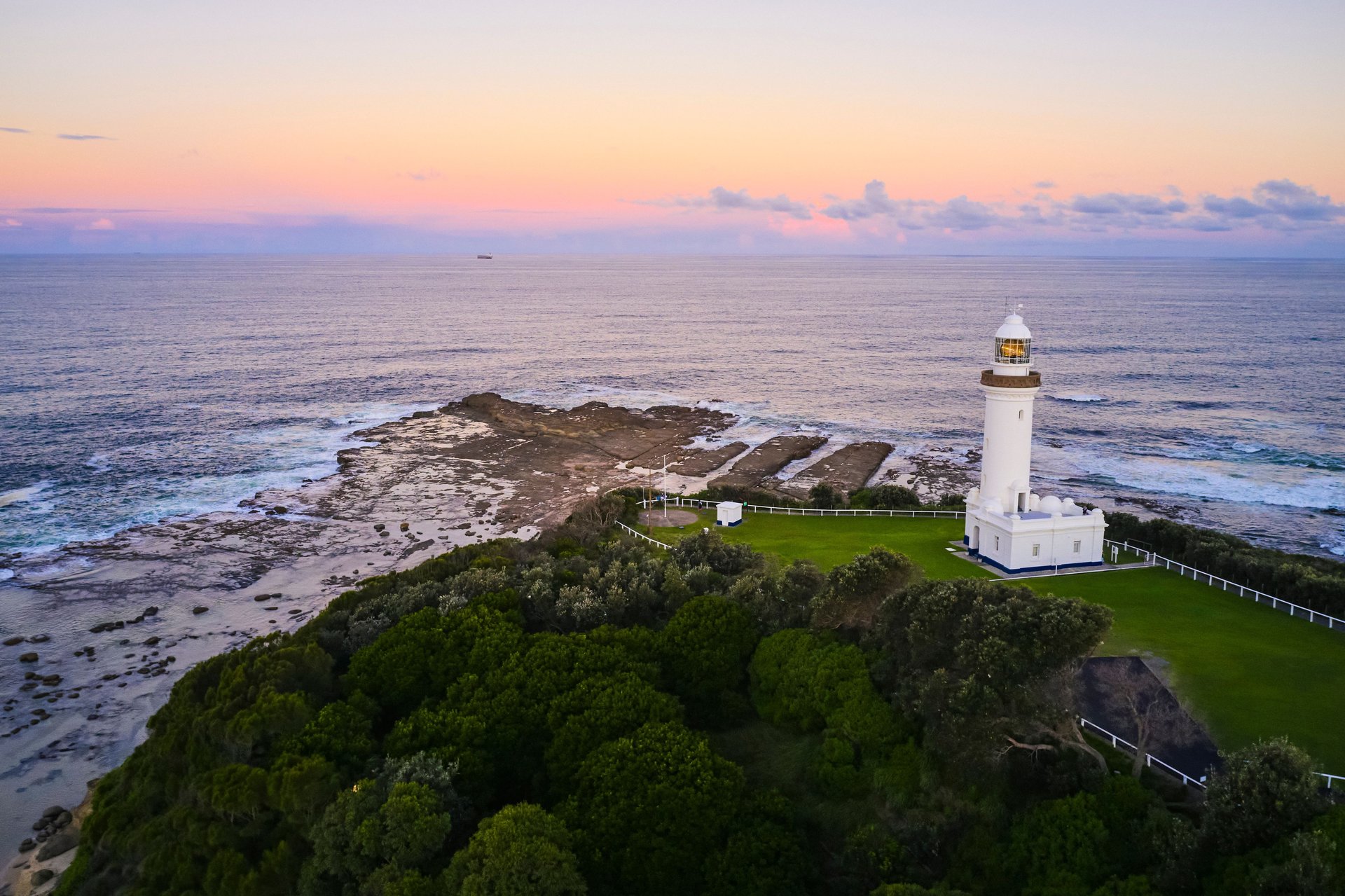 White lighthouse on a green coastal bluff overlooking a rocky shoreline and calm ocean under a pastel sunset sky.