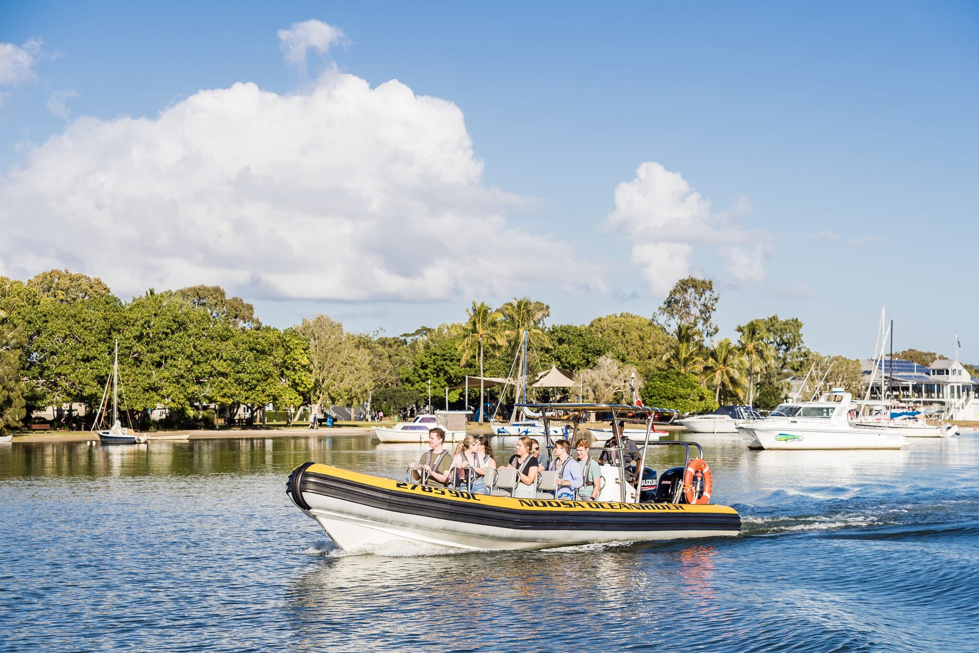 A small boat with passengers cruises on a calm river, surrounded by trees and moored boats under a clear blue sky.
