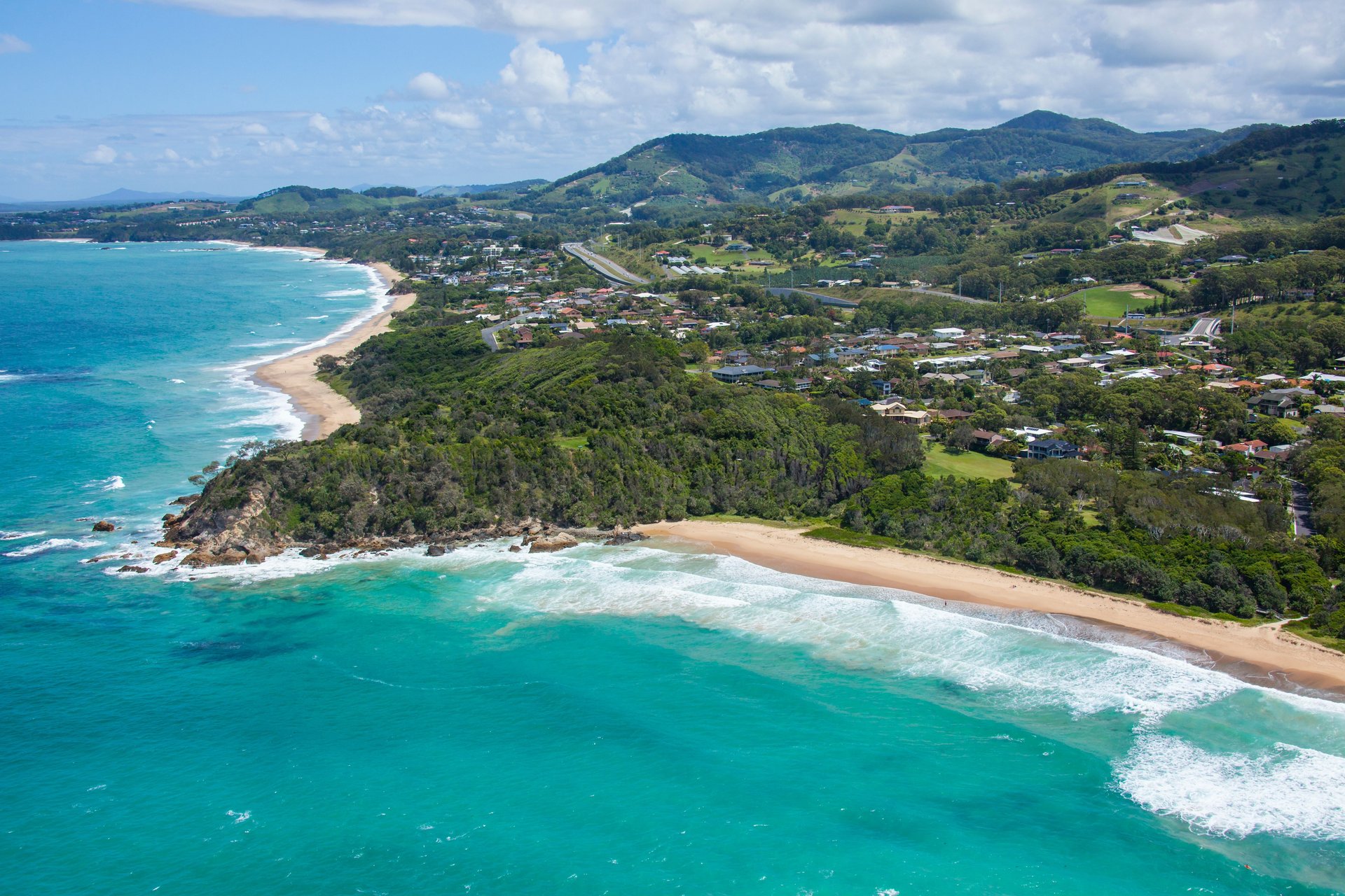 Aerial view of turquoise ocean and sandy beaches curving around a rocky, tree-covered headland with a coastal town and rolling green hills.