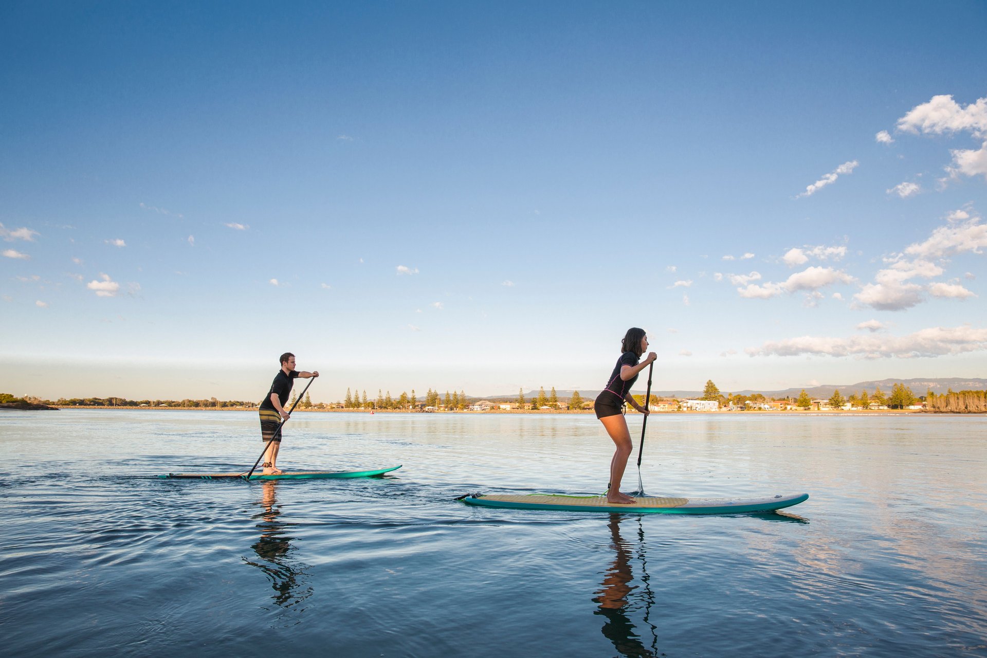 A woman and man stand-up paddleboarding on calm water beneath a clear blue sky with a distant shoreline.