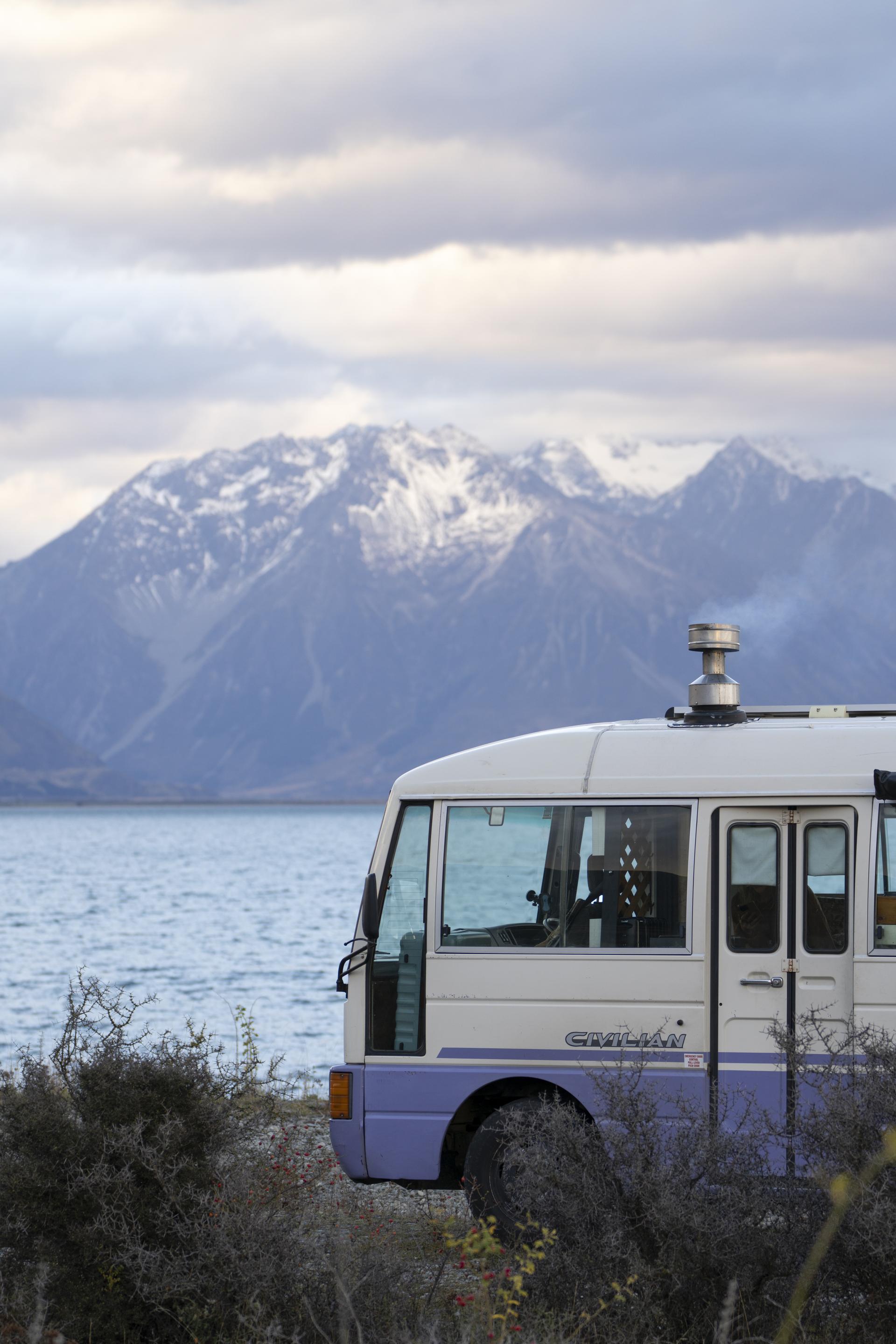 A Camplify white and purple campervan parked at a lake in New Zealand overlooking the mountains