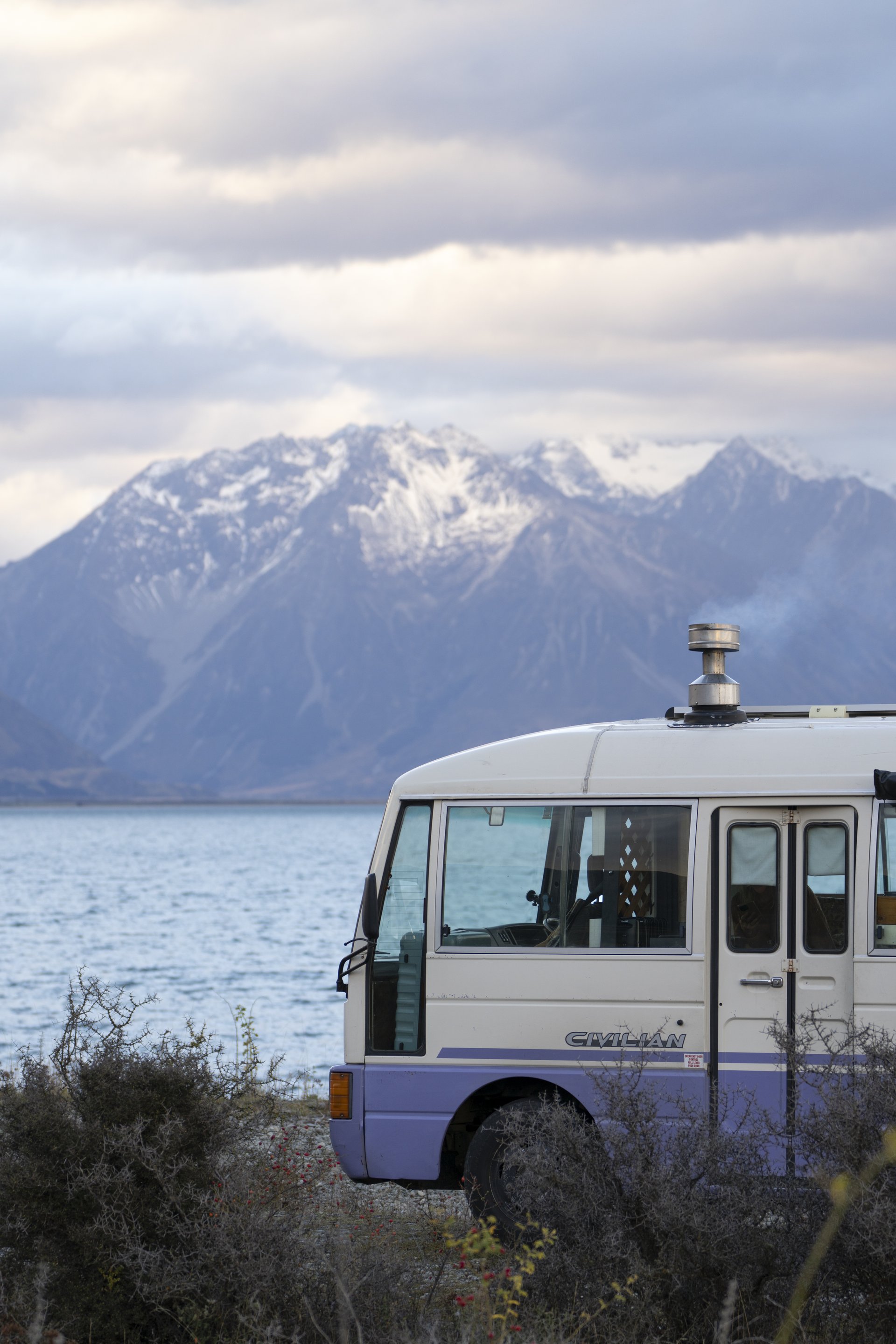 A Camplify white and purple campervan parked at a lake in New Zealand overlooking the mountains