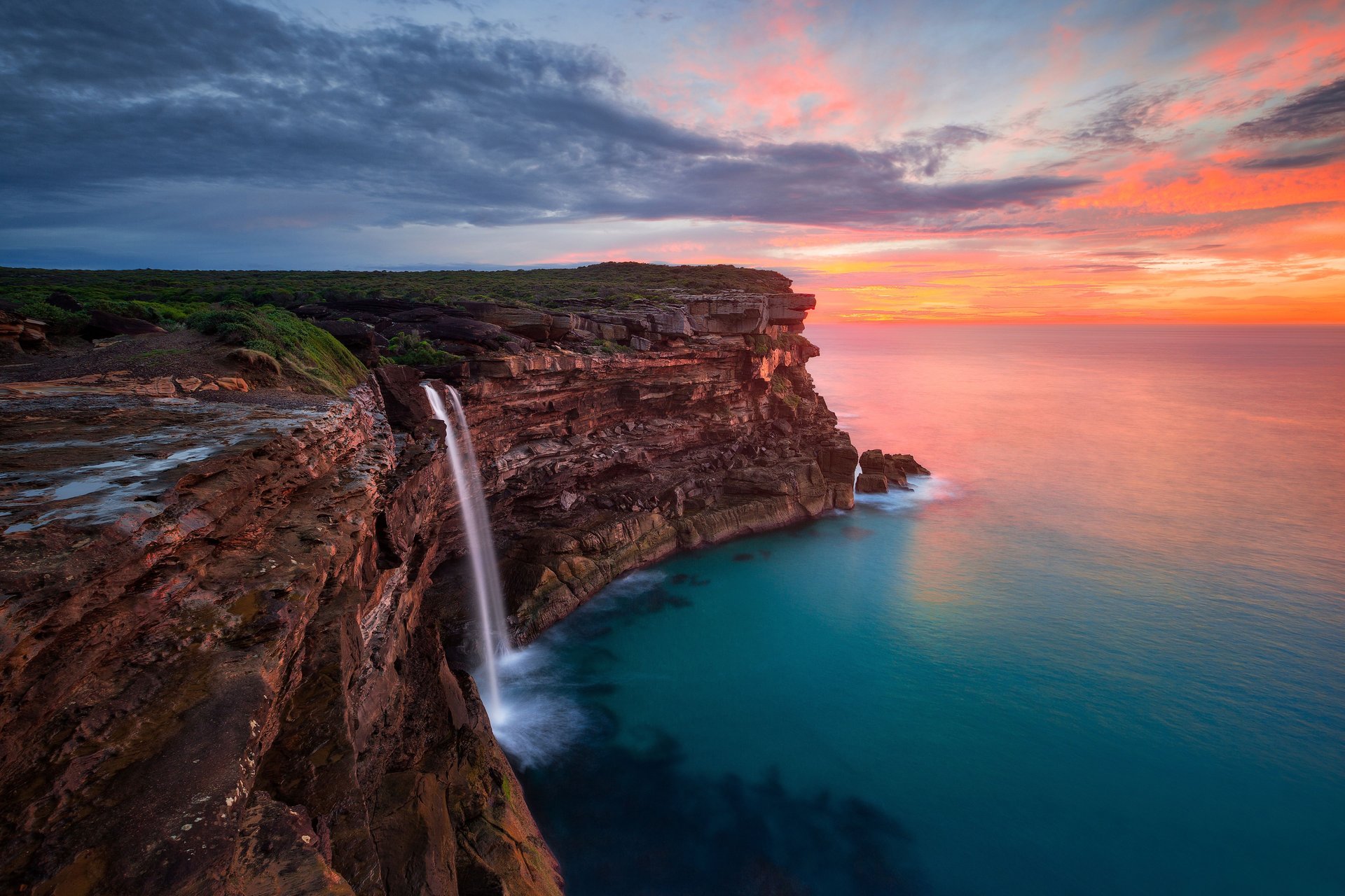 A waterfall cascades off a rocky cliff into a turquoise sea at sunset, with vibrant orange and pink clouds in the sky.