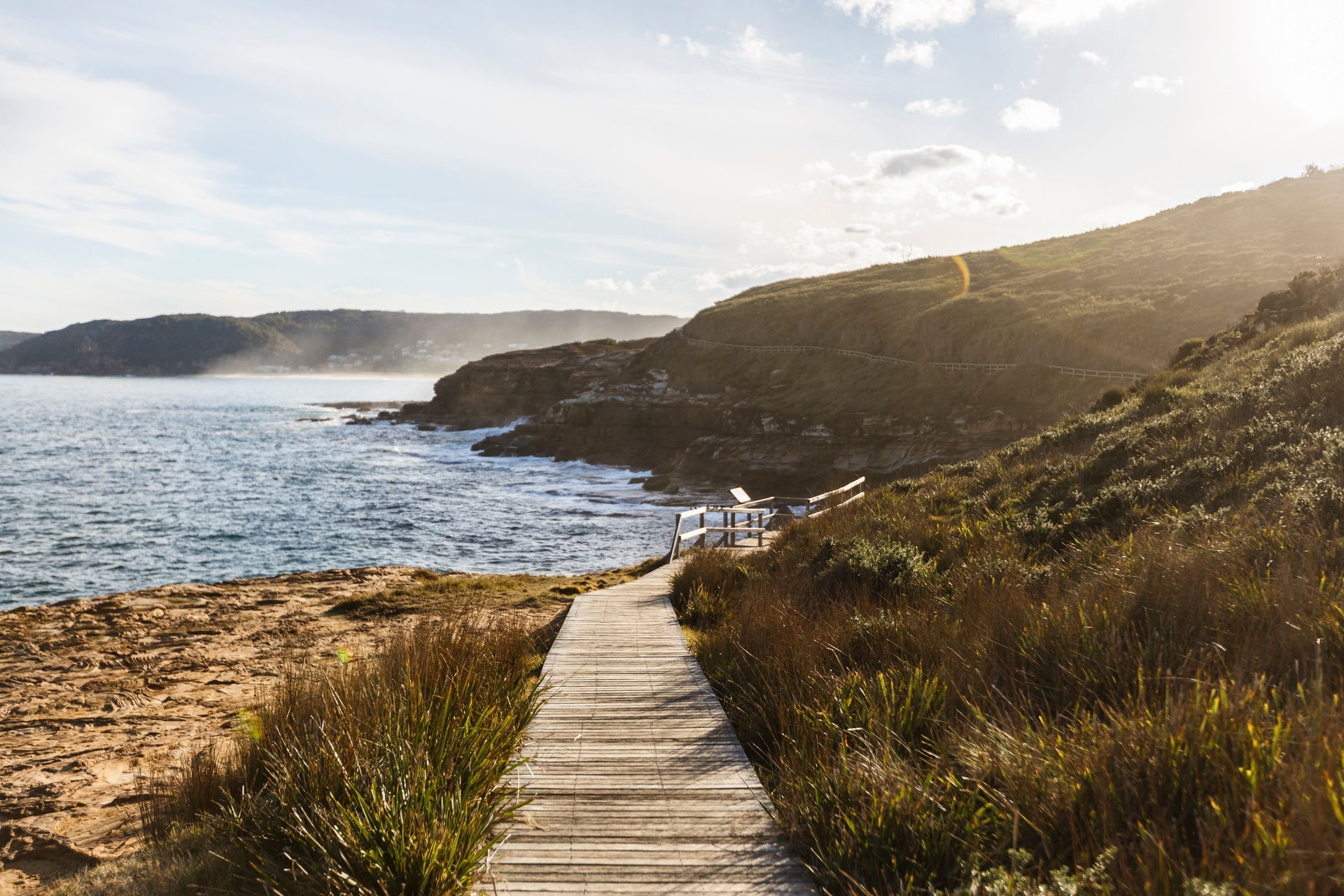 Wooden boardwalk leading to a coastal lookout over rocky cliffs and ocean, bathed in golden sunlight with grassy dunes.