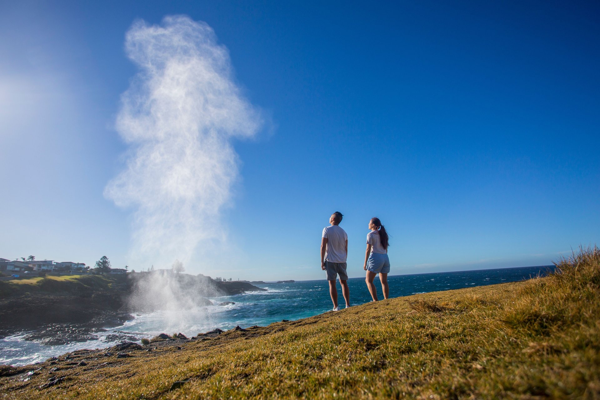Two people on a grassy coastal cliff watching a tall plume of sea spray from a blowhole against a blue sky.