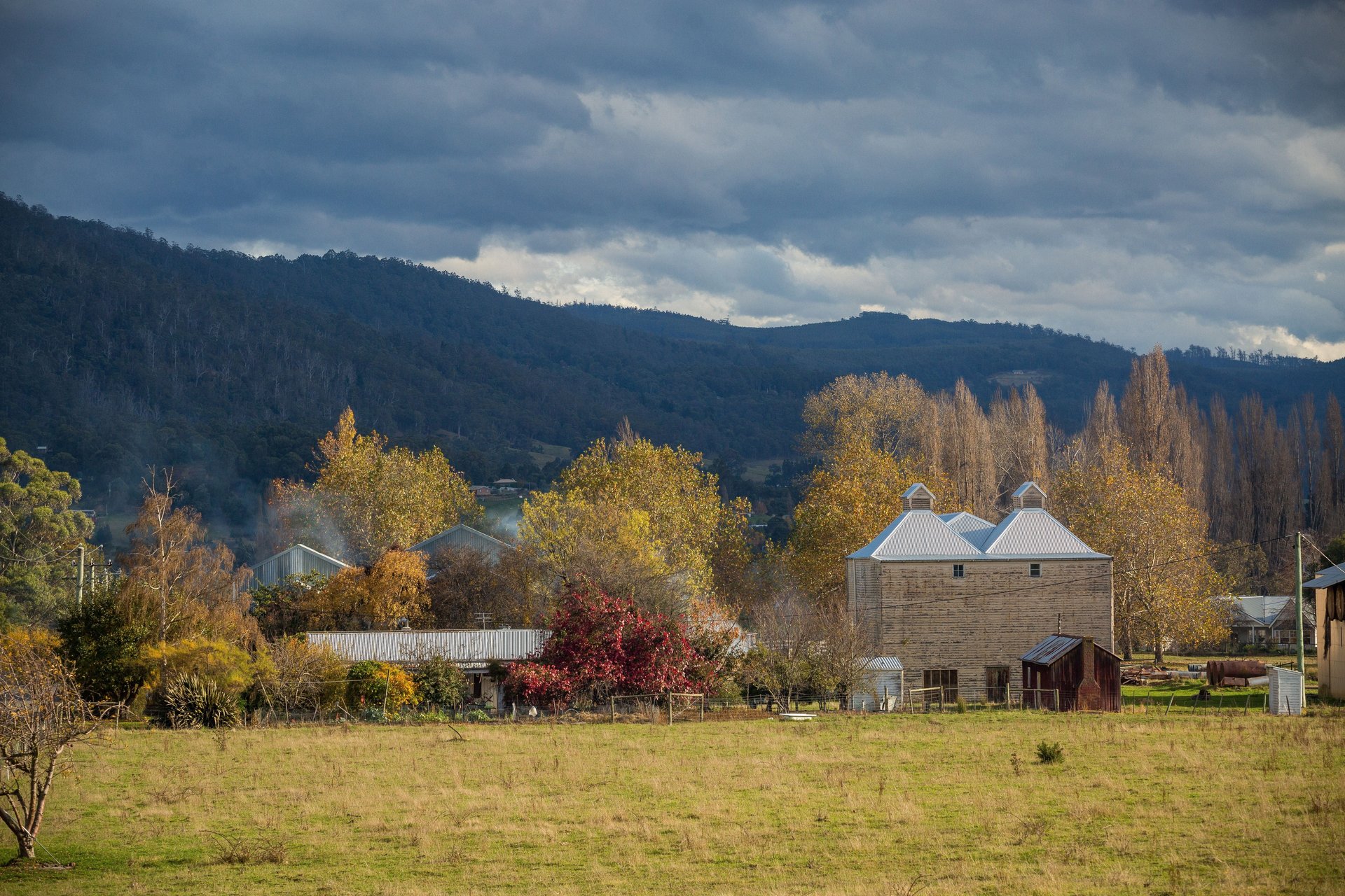 Rural landscape with a stone barn, autumn trees, and distant hills under a cloudy sky.
