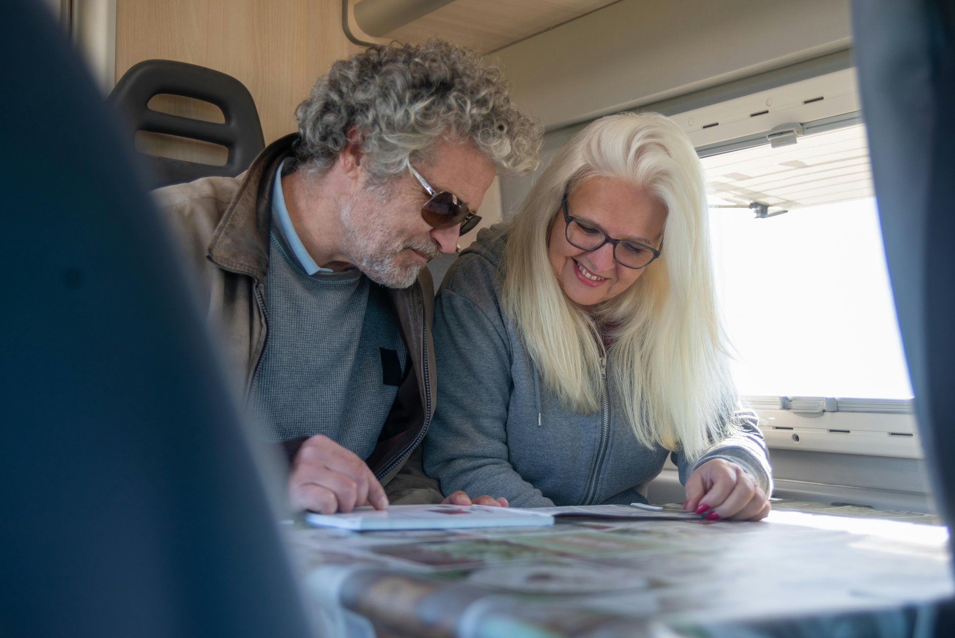 A man and woman sit in a camper van, smiling and looking at a book together. The man wears sunglasses and a jacket; the woman wears glasses.