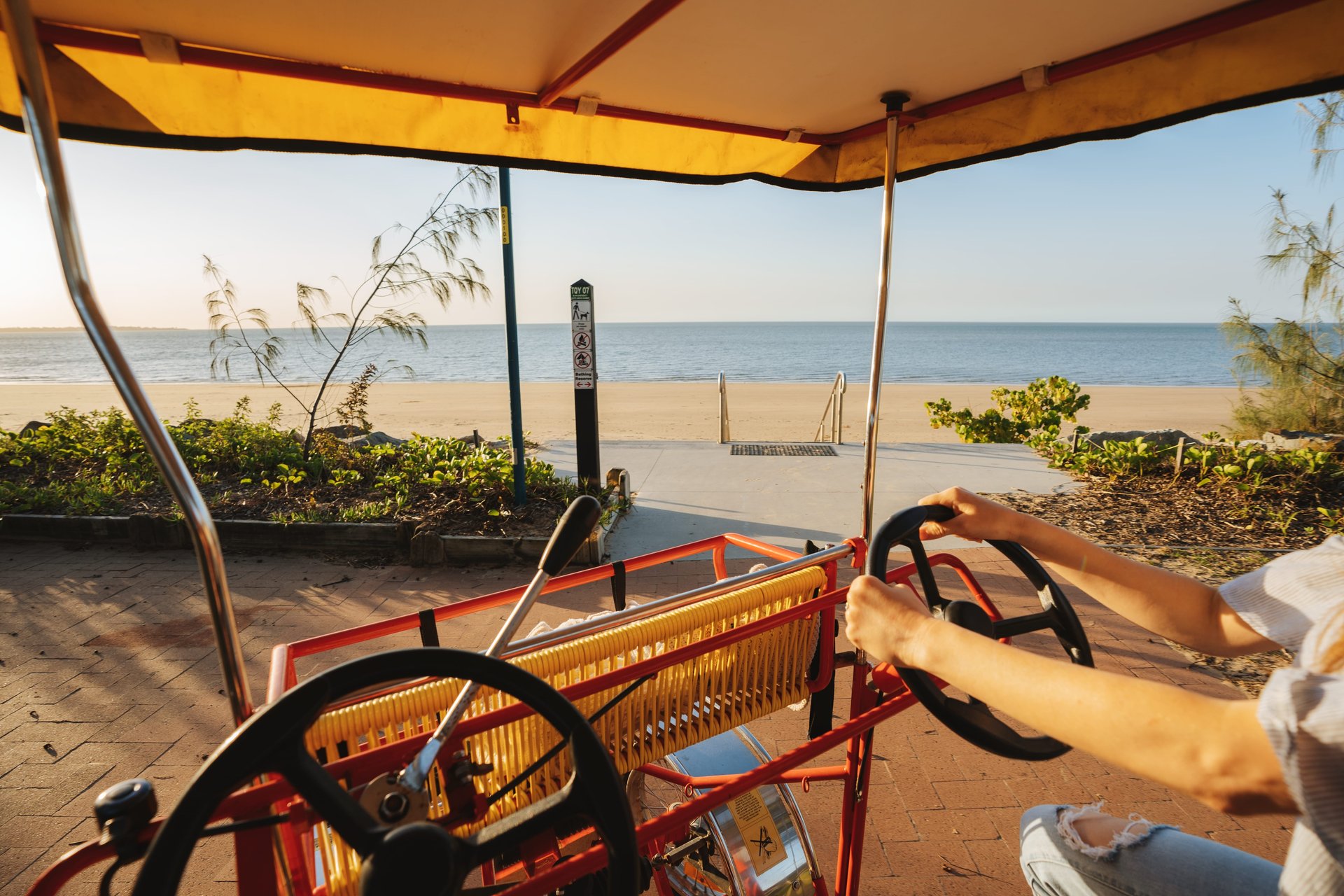 View from a yellow-canopied beach surrey with hands on the wheel, path leading to a sandy beach and calm sea horizon.