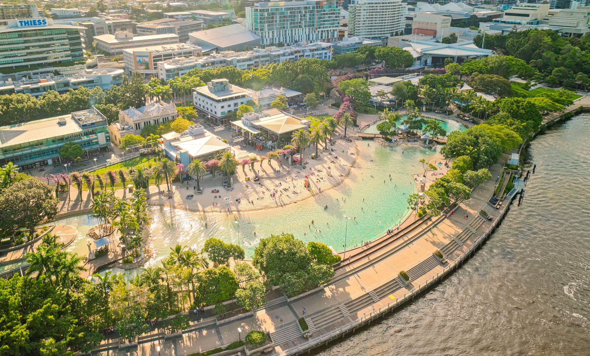 Aerial view of a bustling urban beach with palm trees, surrounded by city buildings and a river, under a clear sky.