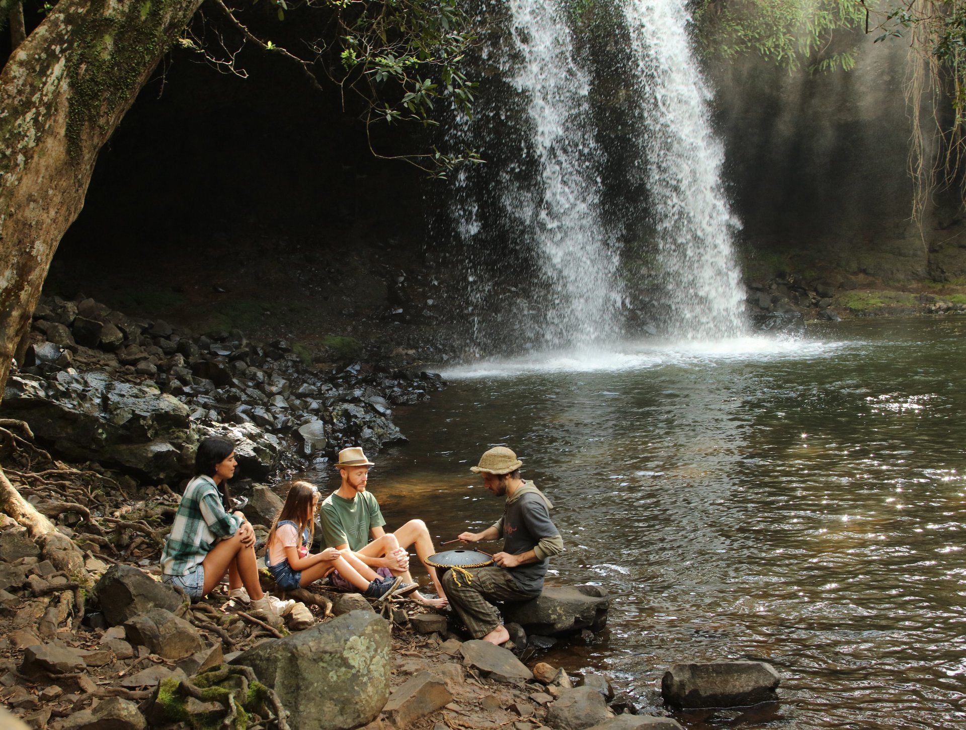 Four people seated on a rocky shore below a waterfall, one person playing a hand drum.