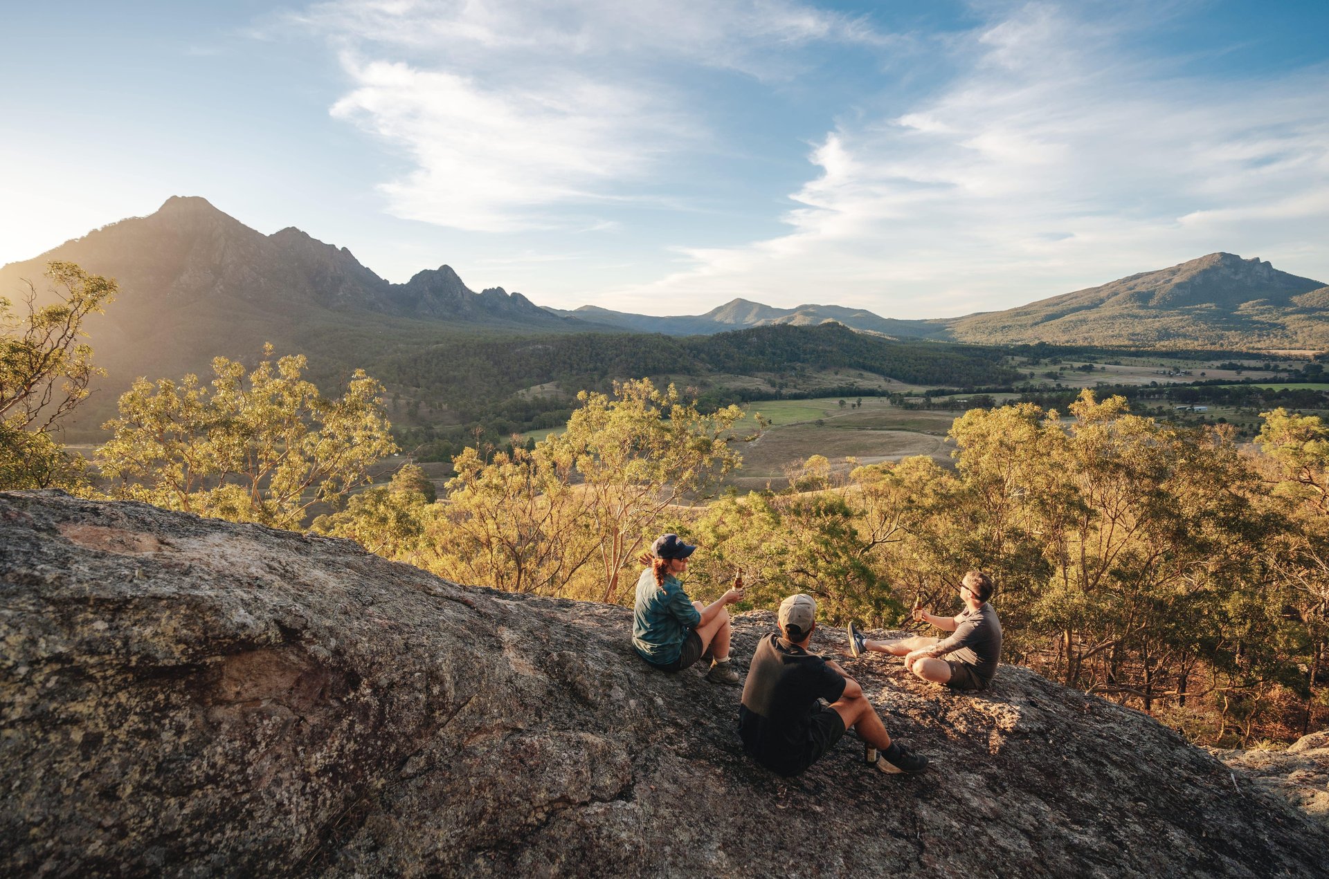 Three people sit on a rocky ledge overlooking a scenic valley with mountains and trees under a partly cloudy sky at sunset.