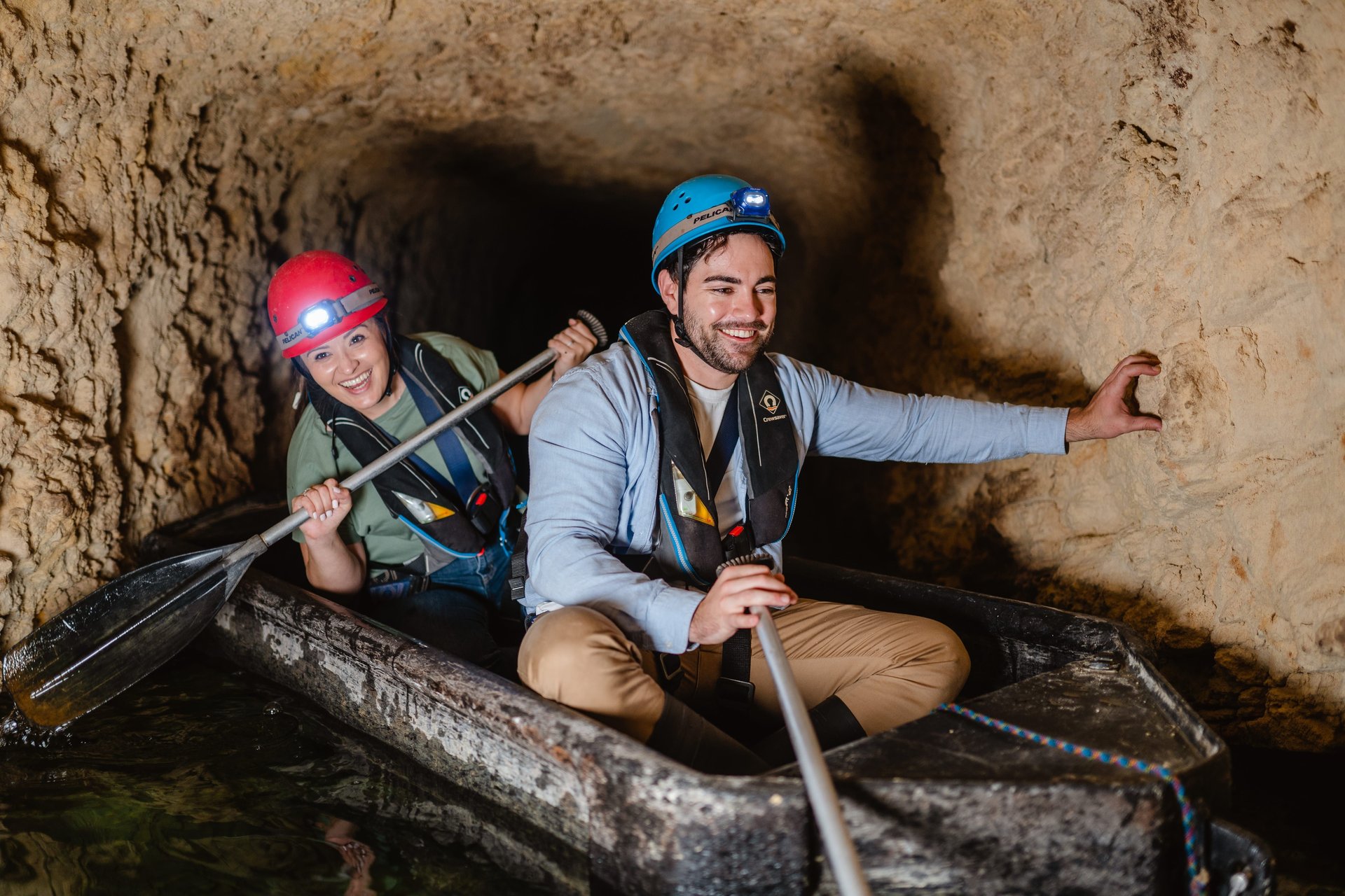 Two people wearing helmets and life vests paddle a small boat through a narrow, rocky cave tunnel, smiling and enjoying the adventure.