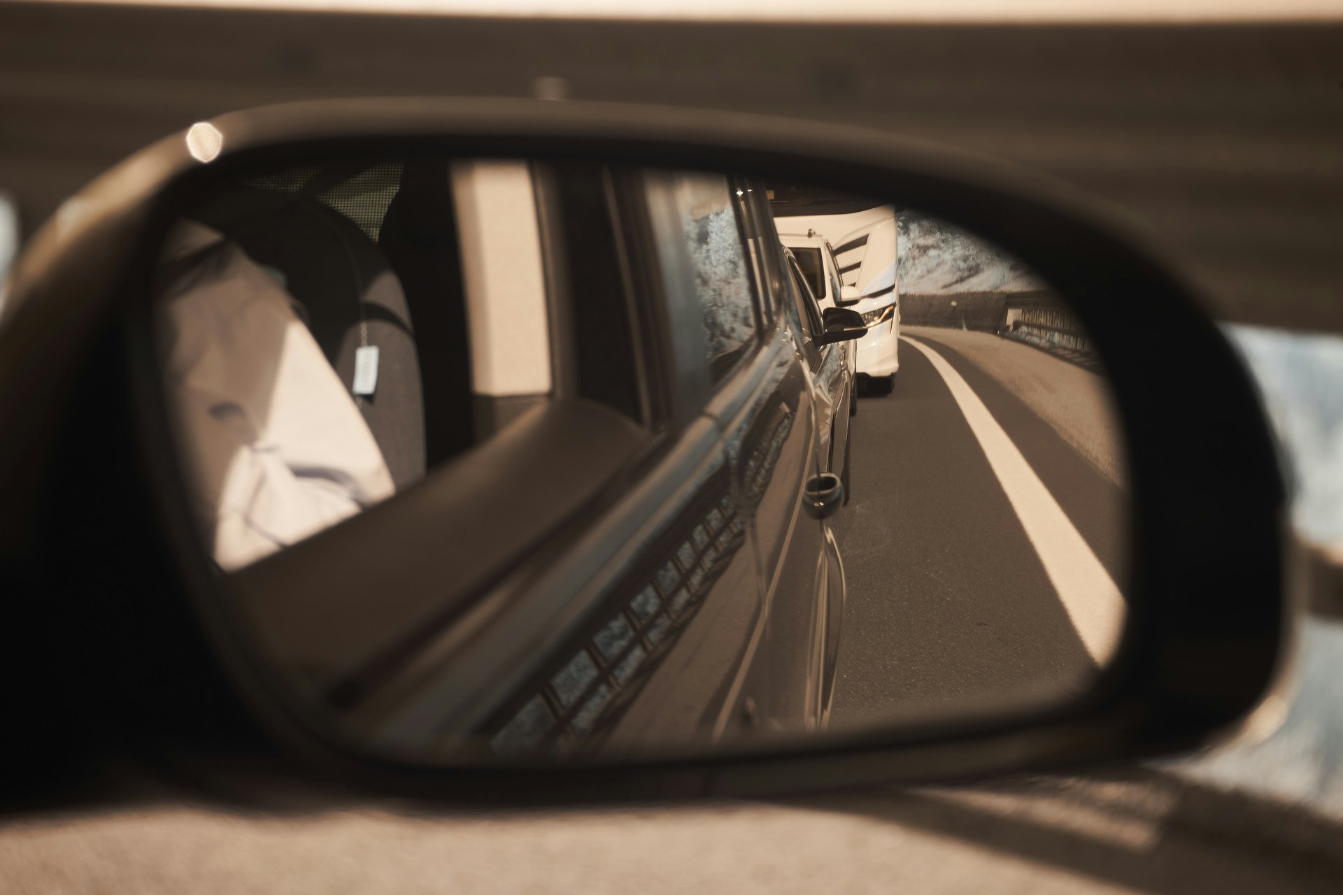 Side mirror view showing a white vehicle behind on a highway, with a blurred guardrail and road markings visible.