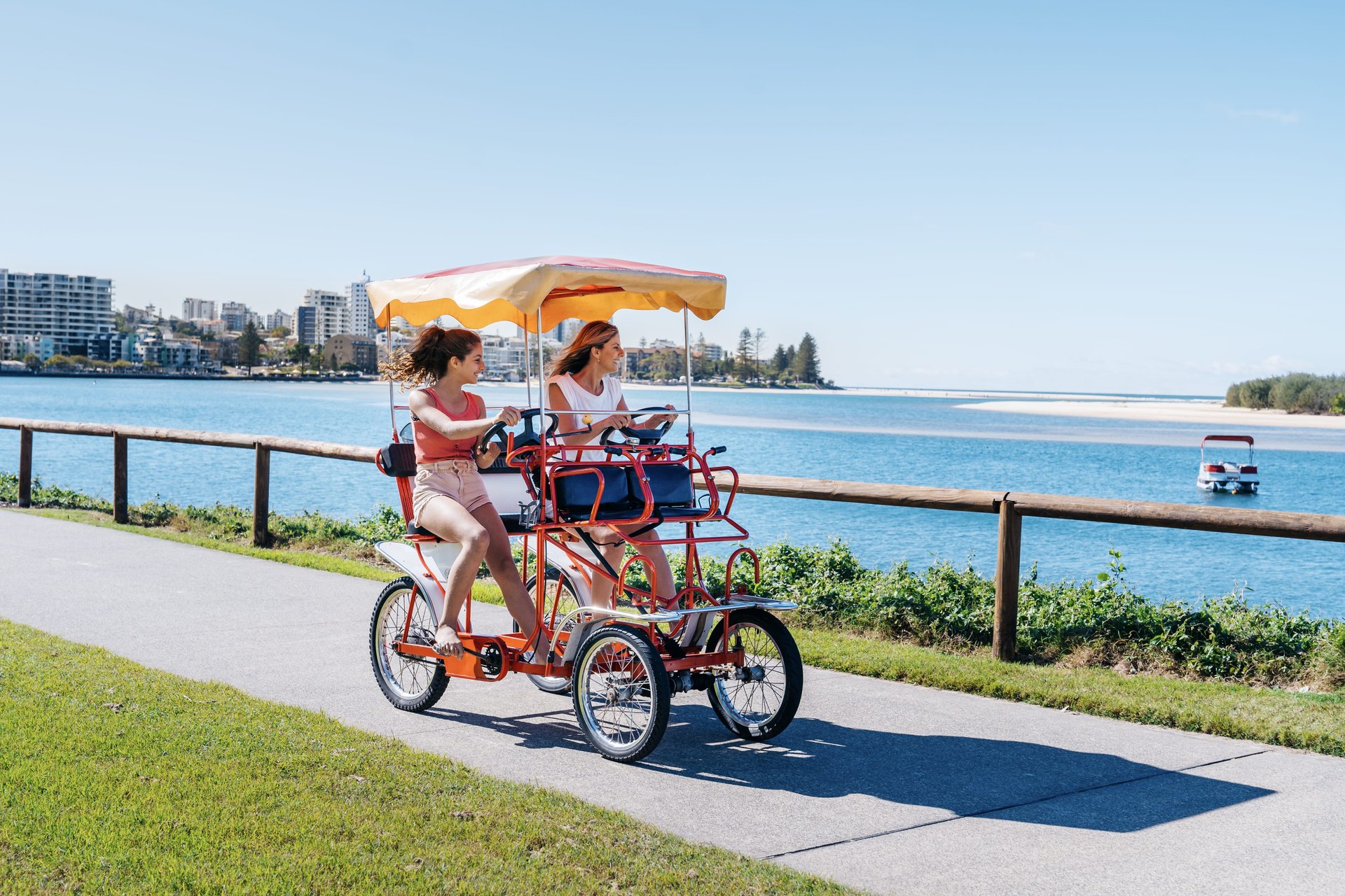 Two people ride a four-wheeled tandem bike with a canopy along a waterfront path on a sunny day, with buildings and a boat in the background.