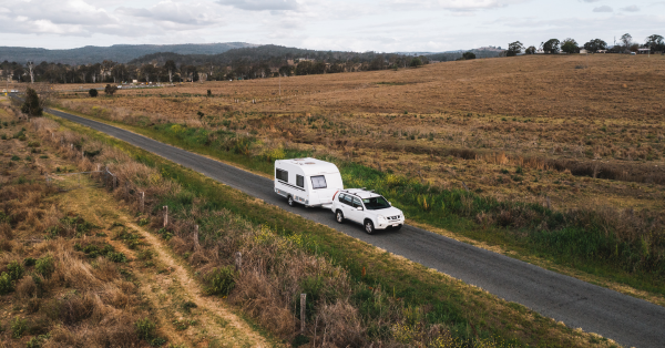 A white car tows a caravan on a narrow rural road, surrounded by open fields and distant hills under a cloudy sky.