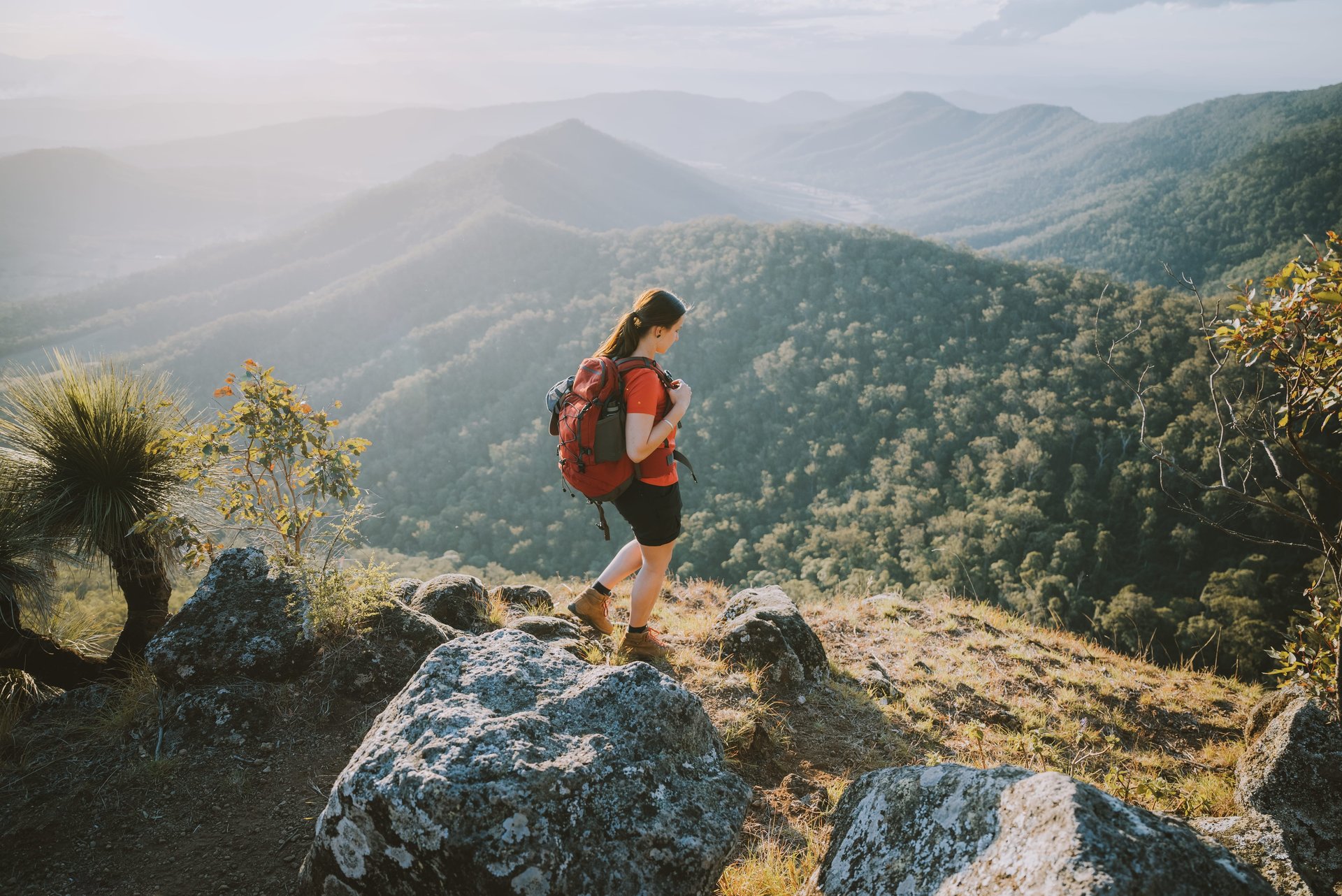 A hiker with a red backpack stands on a rocky ledge, overlooking a vast, mountainous landscape under a clear sky.